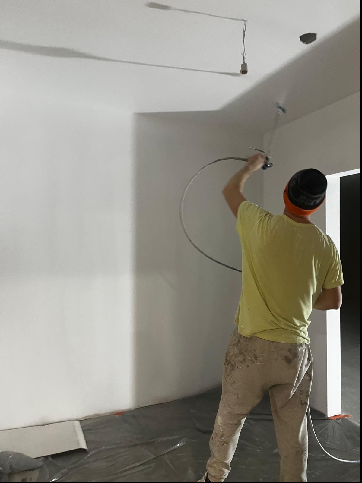 Malowanie ścian natryskowo w pomieszczeniu z odkrytą żarówką, widoczne spodnie malarza z zabrudzeniami farby.