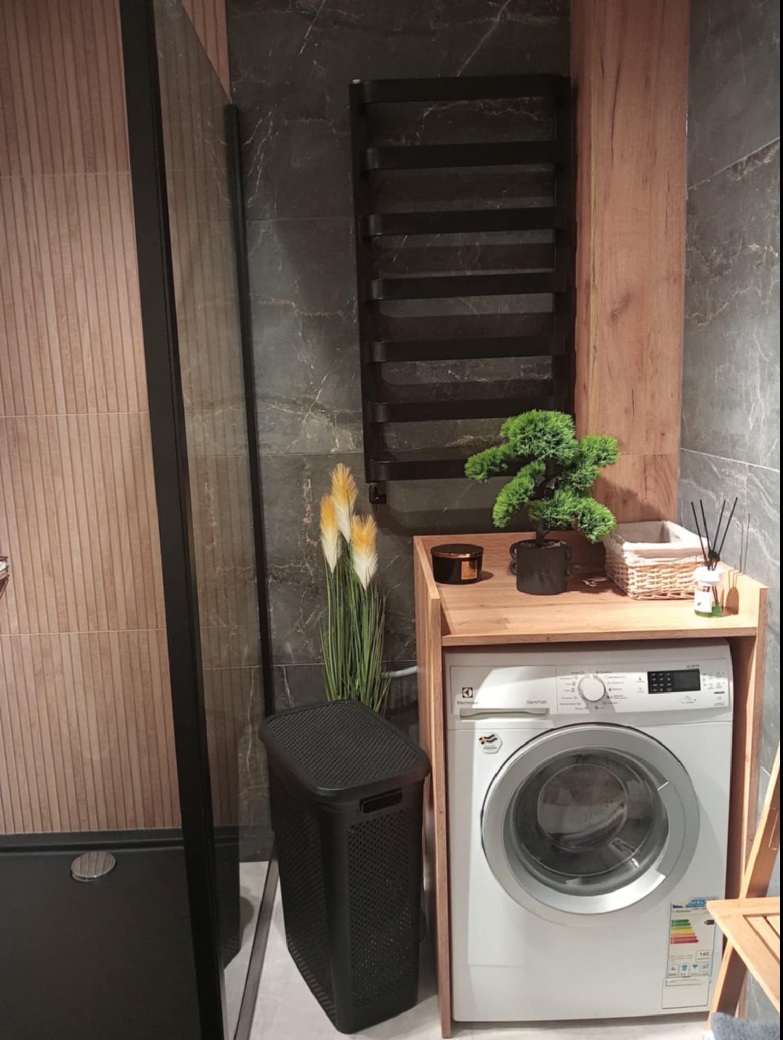 Nowoczesna łazienka z pralką zabudowaną w drewnianej szafce, dekorowana roślinami i czarnym koszem na pranie, obok kabina prysznicowa z ciemną armaturą.