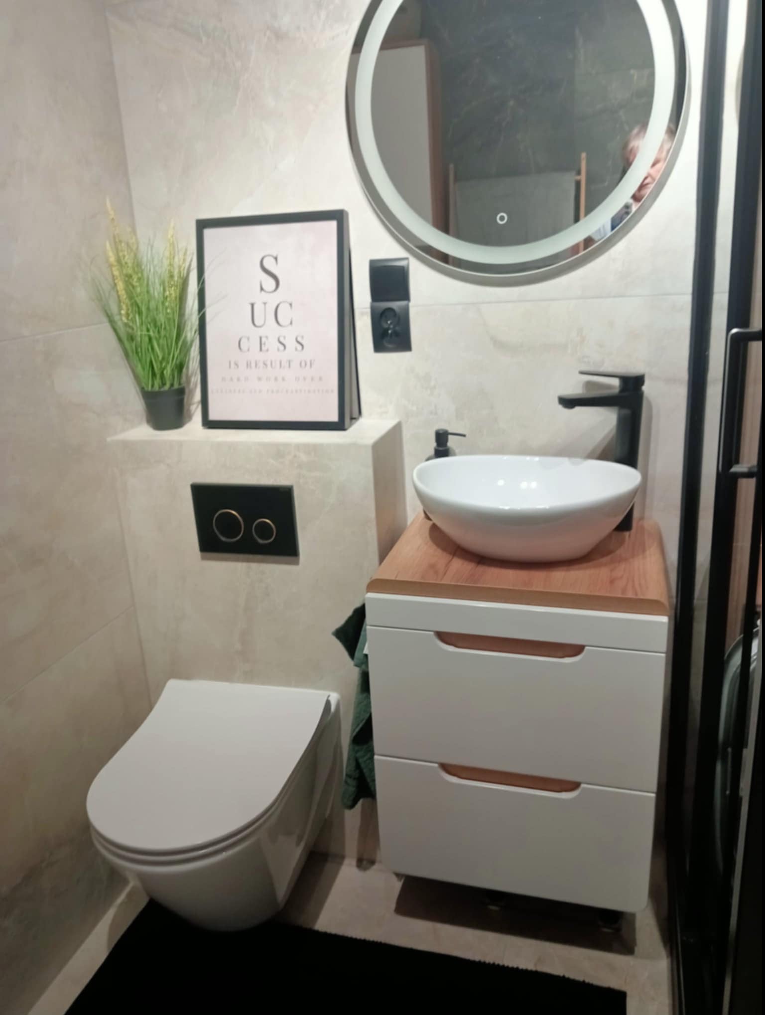 Nowoczesna łazienka w jasnych barwach, z podświetlanym okrągłym lustrem, białą umywalką nablatową na szafce z drewnianym blatem, wiszącą toaletą i dekoracją w postaci ramki z napisem 'Success'.