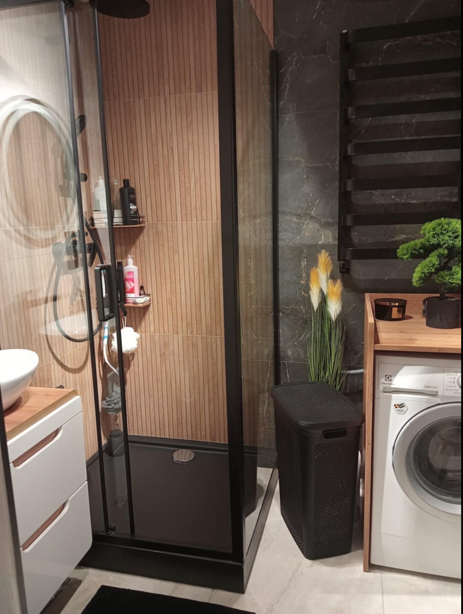 Nowoczesna łazienka z kabiną prysznicową typu walk-in z czarnymi profilami, umywalką nablatową z szafką, pralką Electrolux, grzejnikiem drabinkowym i dekoracyjnymi trawami.