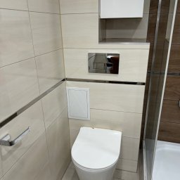 Krzysztof Szewc - Kompaktowa toaleta podwieszana z chromowanym przyciskiem spłukującym, ukryta szafka nad nią, beżowe płytki z metalowymi listwami dekoracyjnymi, widoczny fragment kabiny prysznicowej z brązowymi...