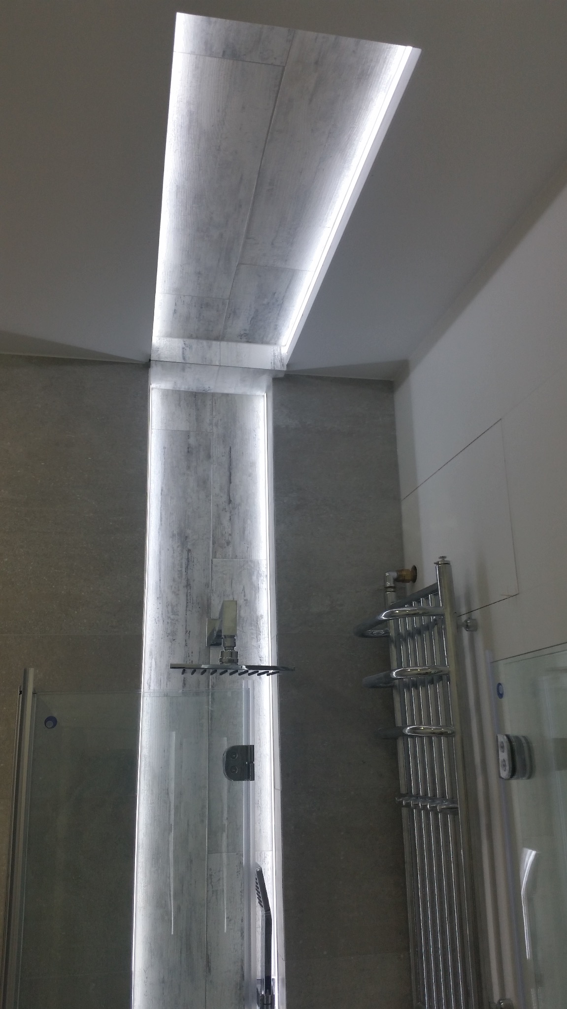 Nowoczesna łazienka z podświetlanym prysznicem typu walk-in i grzejnikiem drabinkowym, wykończona płytkami w odcieniach szarości.