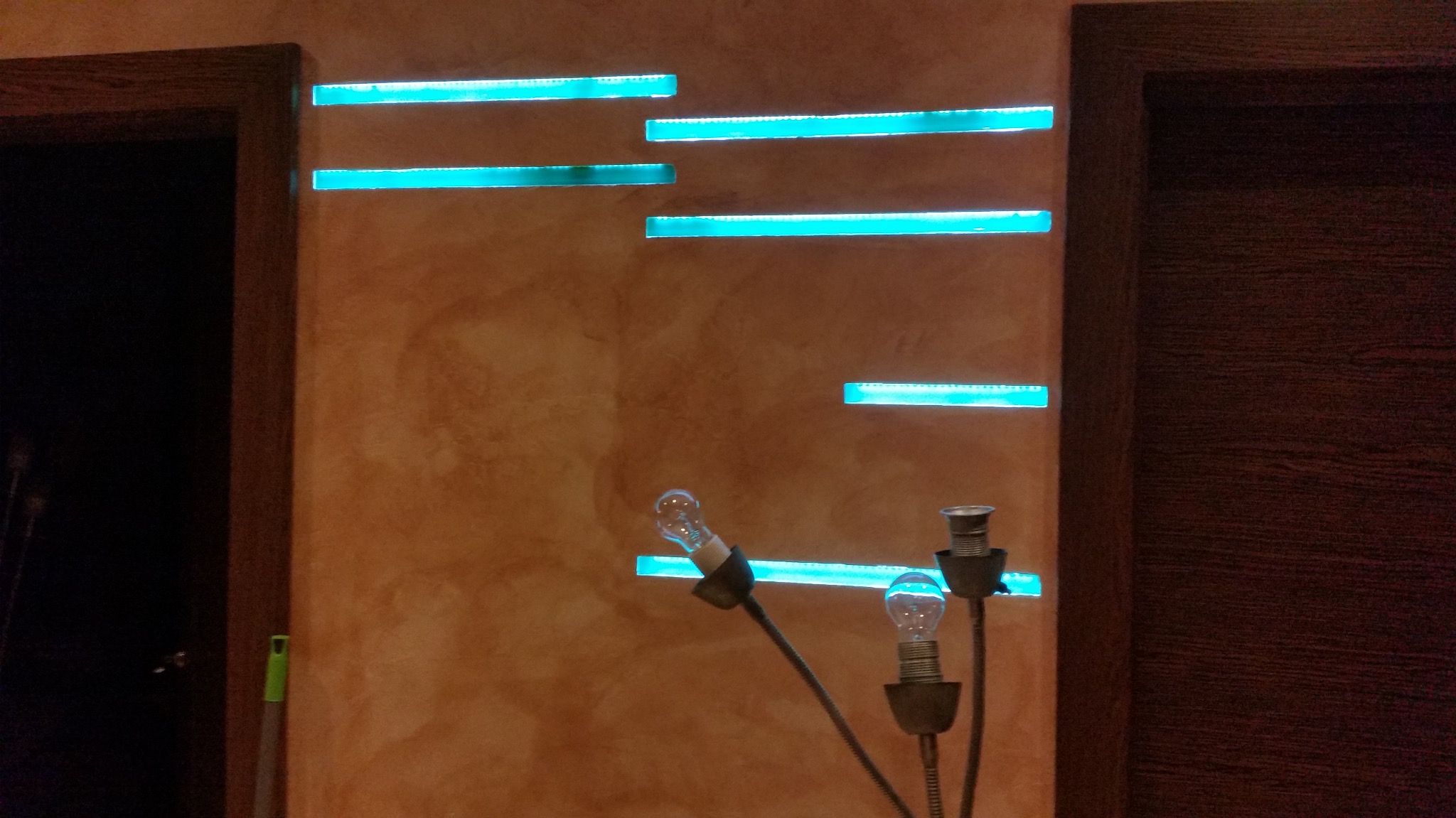 Pomarańczowa ściana z poziomymi wnękami podświetlonymi na niebiesko, widoczna lampa z żarówkami i fragment ramy drzwi.