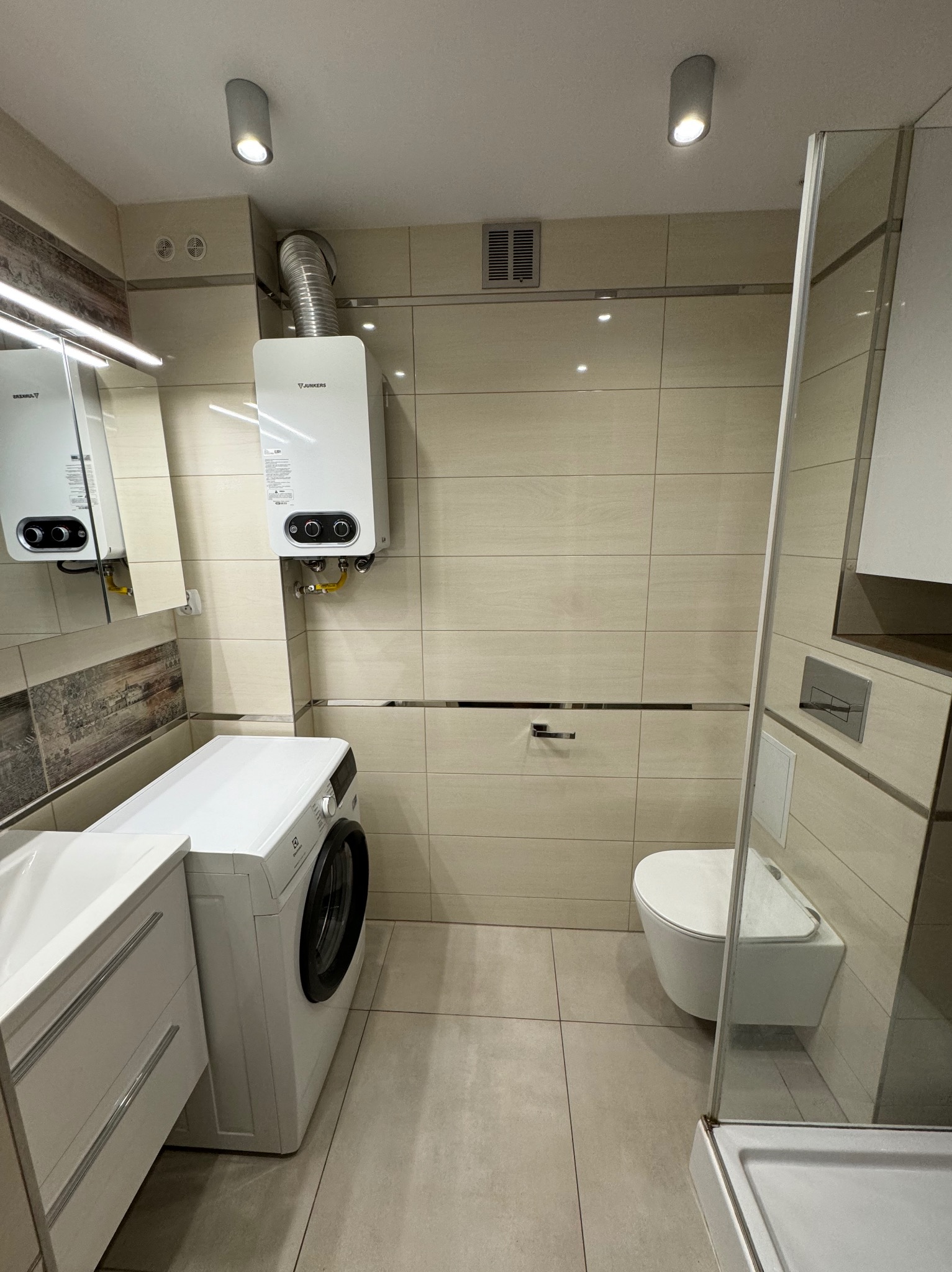 Beżowa łazienka po remoncie: wisząca toaleta, pralka Electrolux, biały bojler Junkers z rurą odprowadzającą spaliny, kabina prysznicowa ze szklaną ścianą, umywalka z szafką i lustrem.