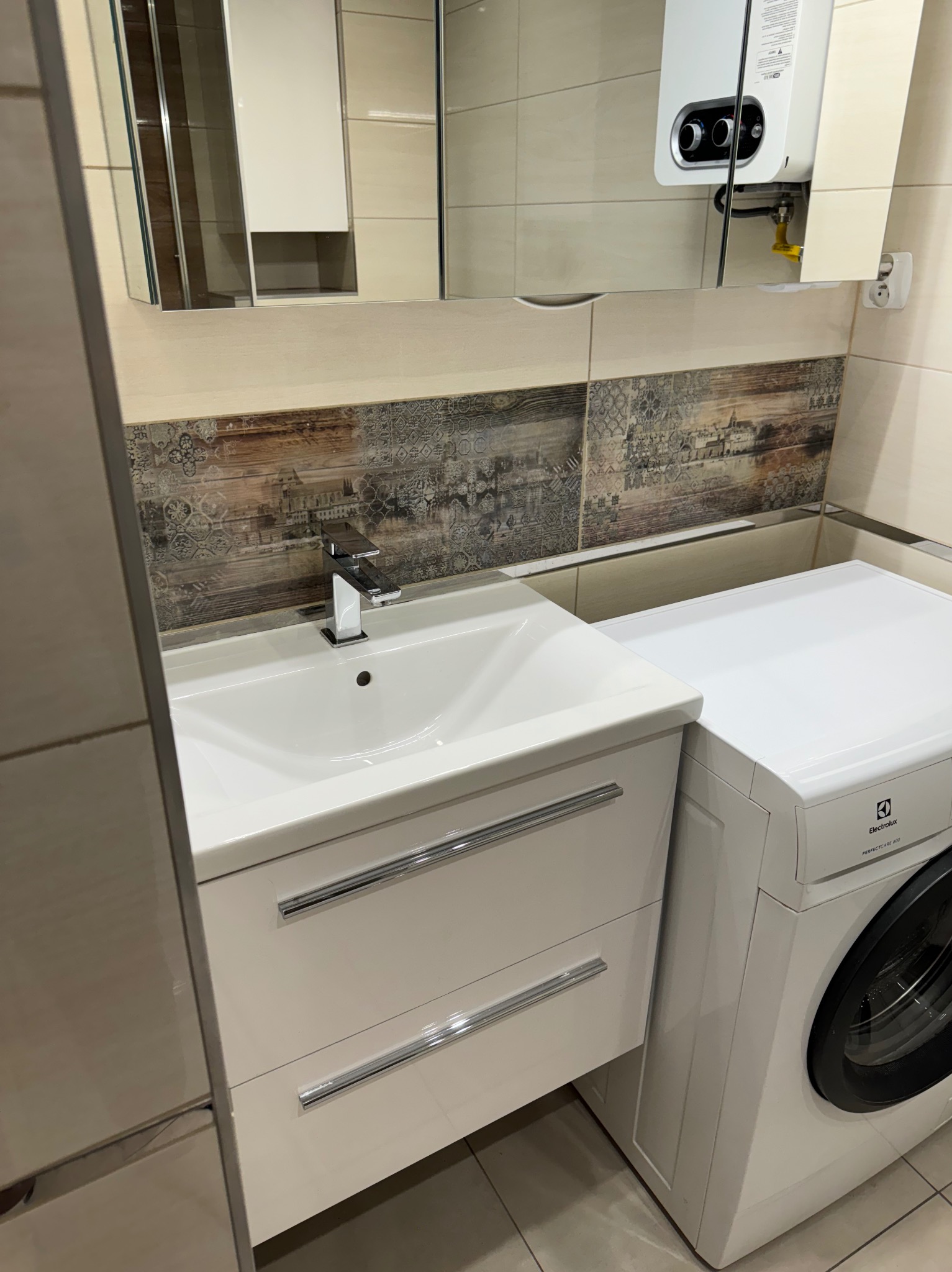 Biała umywalka z szafką, bateria chromowana, pralka Electrolux PerfectCare 600 i dekoracyjne płytki ścienne z motywem miasta w wykończonej łazience.