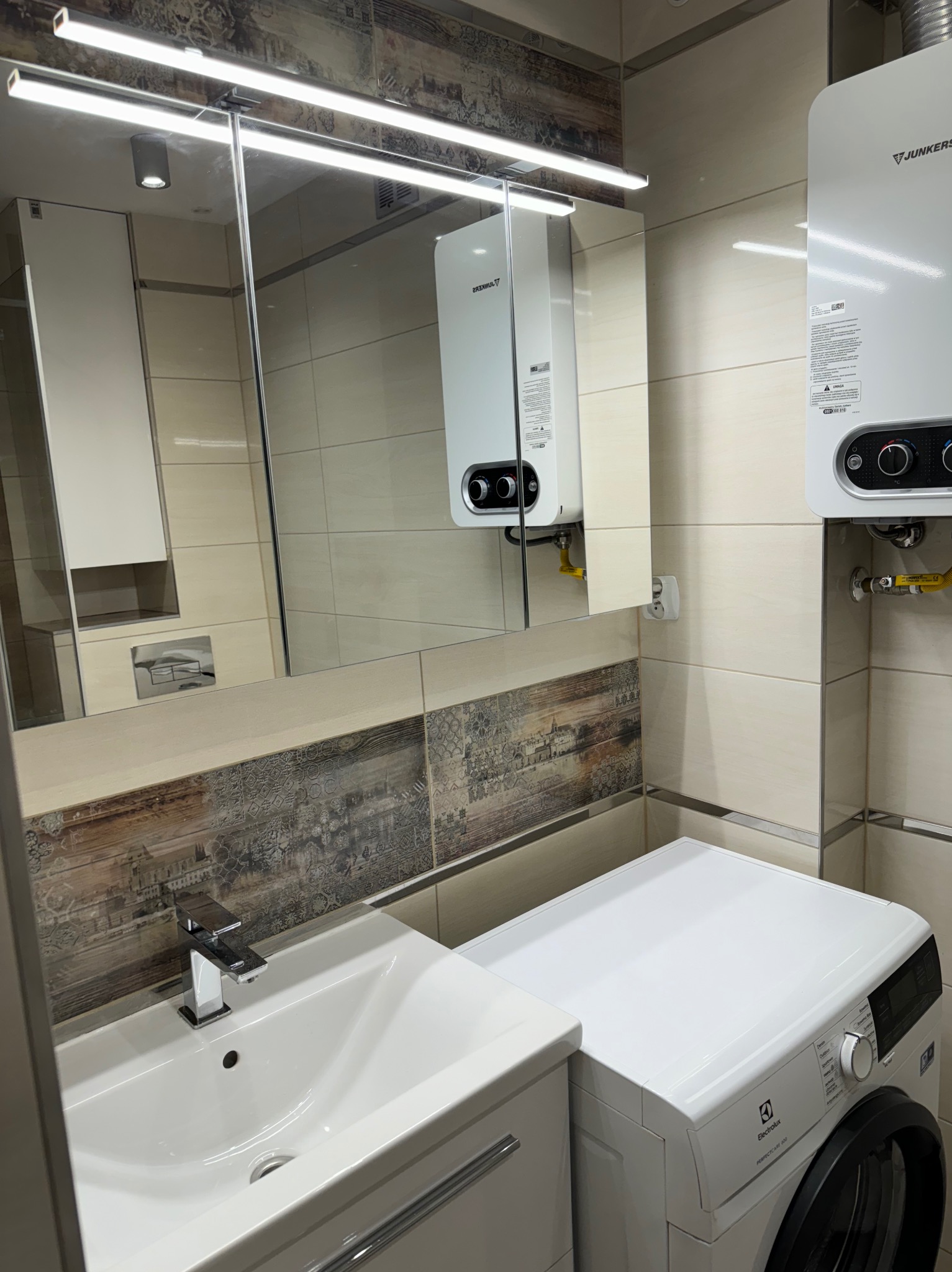 Umywalka z szafką, pralka Electrolux i wiszący piec gazowy Junkers w wykończonej łazience z beżowymi płytkami i dekoracyjnym pasem z motywem miejskim.