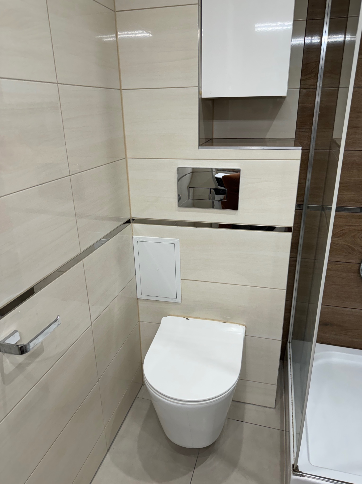 Kompaktowa toaleta podwieszana z chromowanym przyciskiem spłukującym, ukryta szafka nad nią, beżowe płytki z metalowymi listwami dekoracyjnymi, widoczny fragment kabiny prysznicowej z brązowymi...