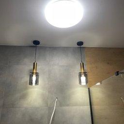 Ecodynamic - Dwa wiszące, szklane klosze z elementami w kolorze złota, oświetlające szarą, kafelkową ścianę; widoczny kabel wychodzący ze ściany i okrągła lampa sufitowa.