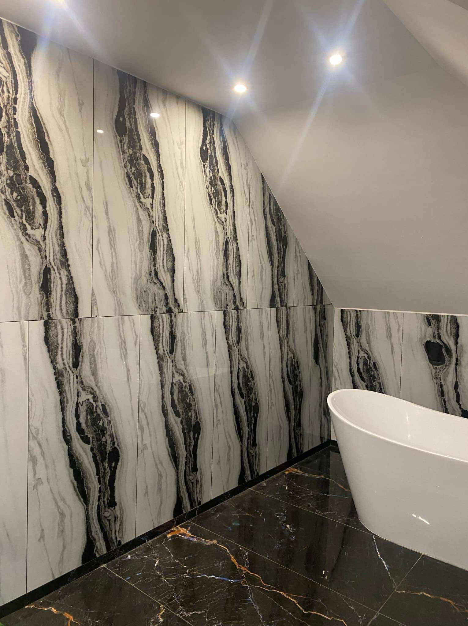 Wykończona łazienka na poddaszu z wanną wolnostojącą, ściany wyłożone płytkami imitującymi marmur z ciemnymi żyłami, podłoga z czarnego marmuru z złotymi żyłami, oświetlenie punktowe.