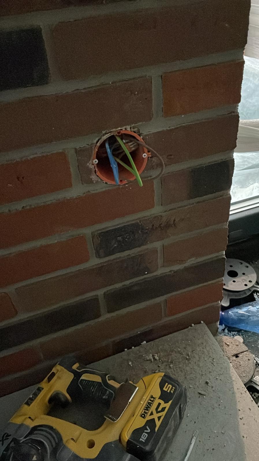 Pomarańczowa puszka elektryczna w ścianie z cegły, widoczne przewody: niebieski, zielony i brązowy, obok leży wiertarko-wkrętarka DeWalt.
