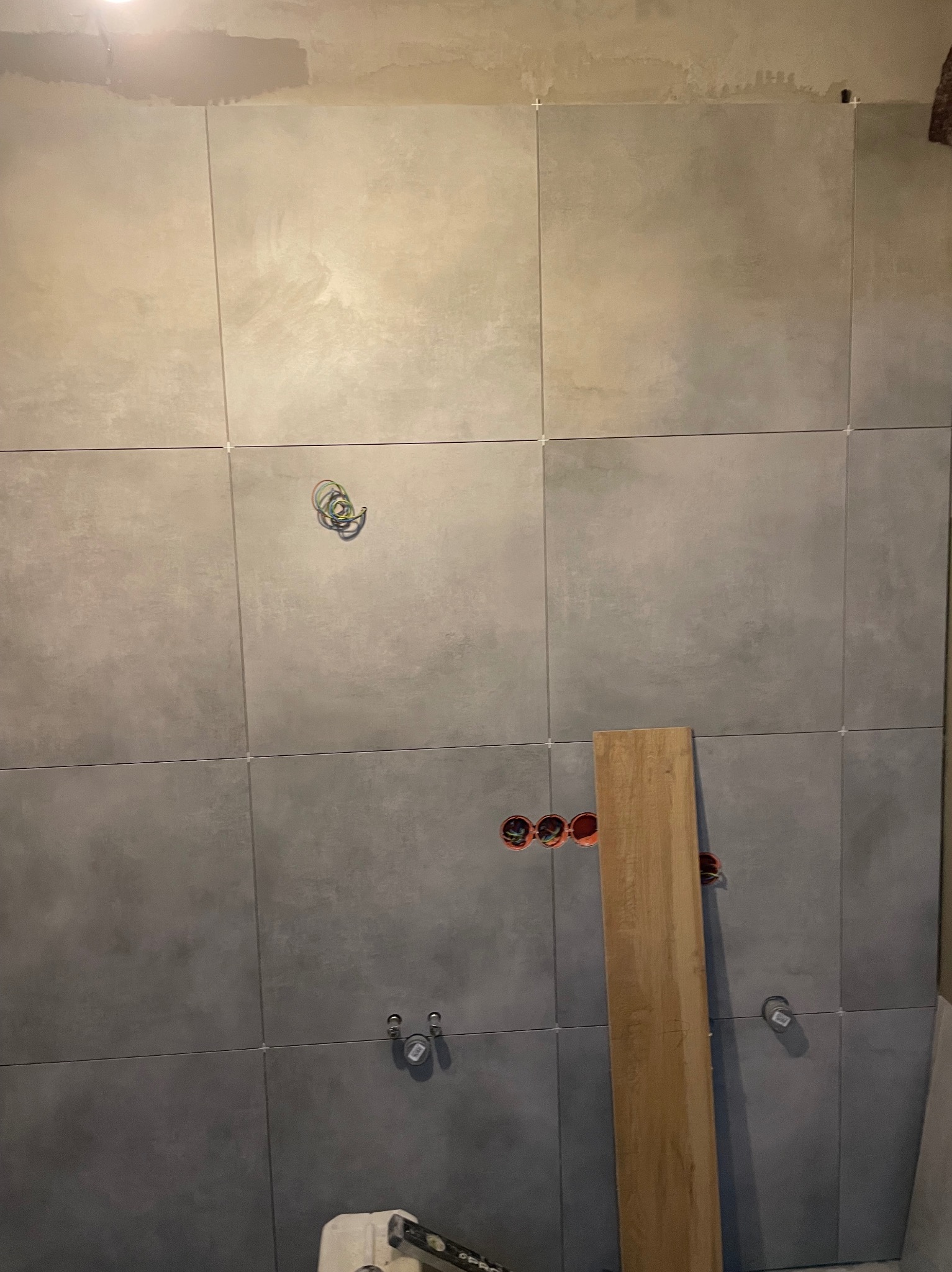 Ściana w trakcie układania płytek ceramicznych w odcieniach szarości, widoczne krzyżyki dystansowe, otwory pod gniazdka elektryczne z pomarańczowymi puszkami, wystające kable oraz przygotowane...