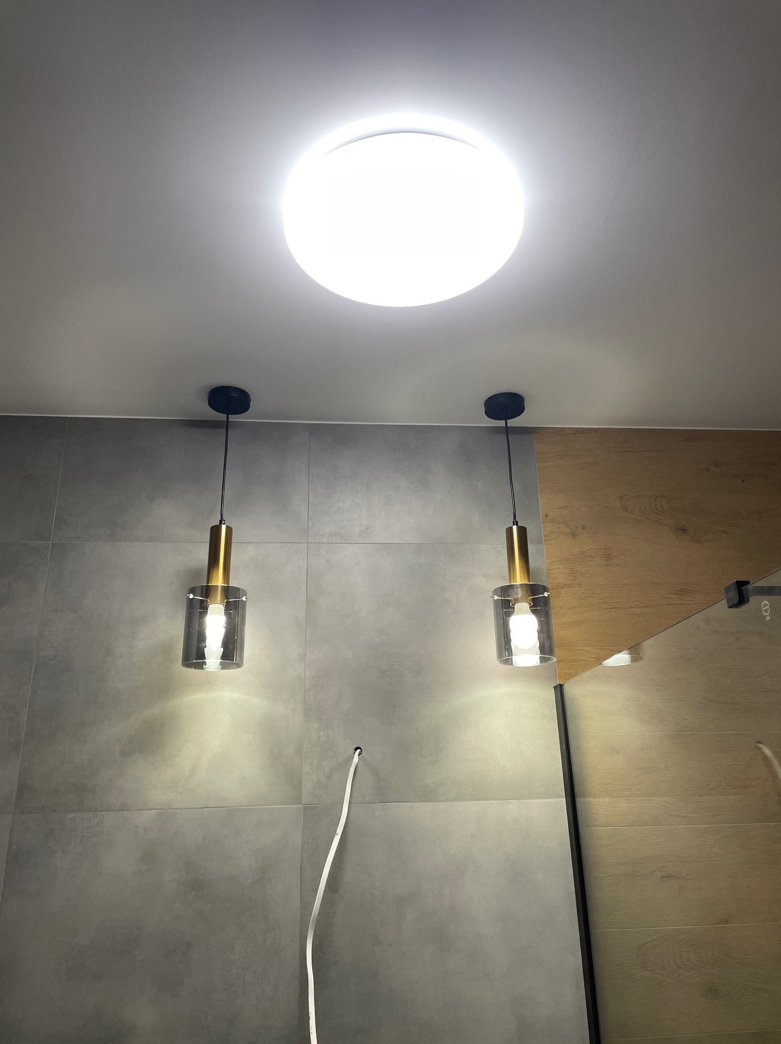 Dwa wiszące, szklane klosze z elementami w kolorze złota, oświetlające szarą, kafelkową ścianę; widoczny kabel wychodzący ze ściany i okrągła lampa sufitowa.