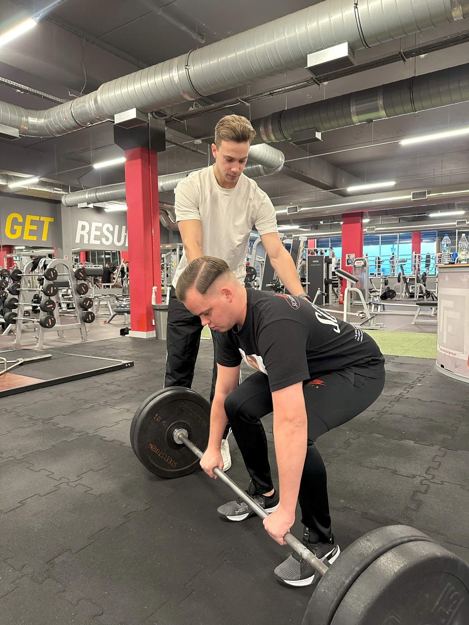 Mężczyzna w czarnej koszulce wykonuje martwy ciąg z asekuracją trenera w białej koszulce, w tle widoczne wyposażenie siłowni i czerwony filar.