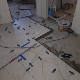 PATRYK KORZBOG-ŁĄCKI - Układanie płytek podłogowych z użyciem poziomicy BMI, krzyżyków dystansowych i taśmy malarskiej; widoczny klej, kielnia i młotek gumowy.