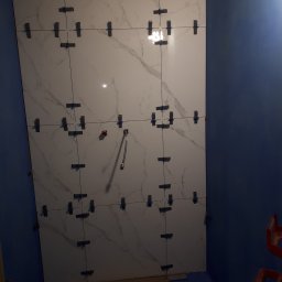 PATRYK KORZBOG-ŁĄCKI - Ściana łazienki w trakcie układania płytek imitujących marmur, z widocznymi krzyżykami dystansowymi i wystającą rurką instalacyjną, niebieskie ściany i podłoga zabezpieczona folią.