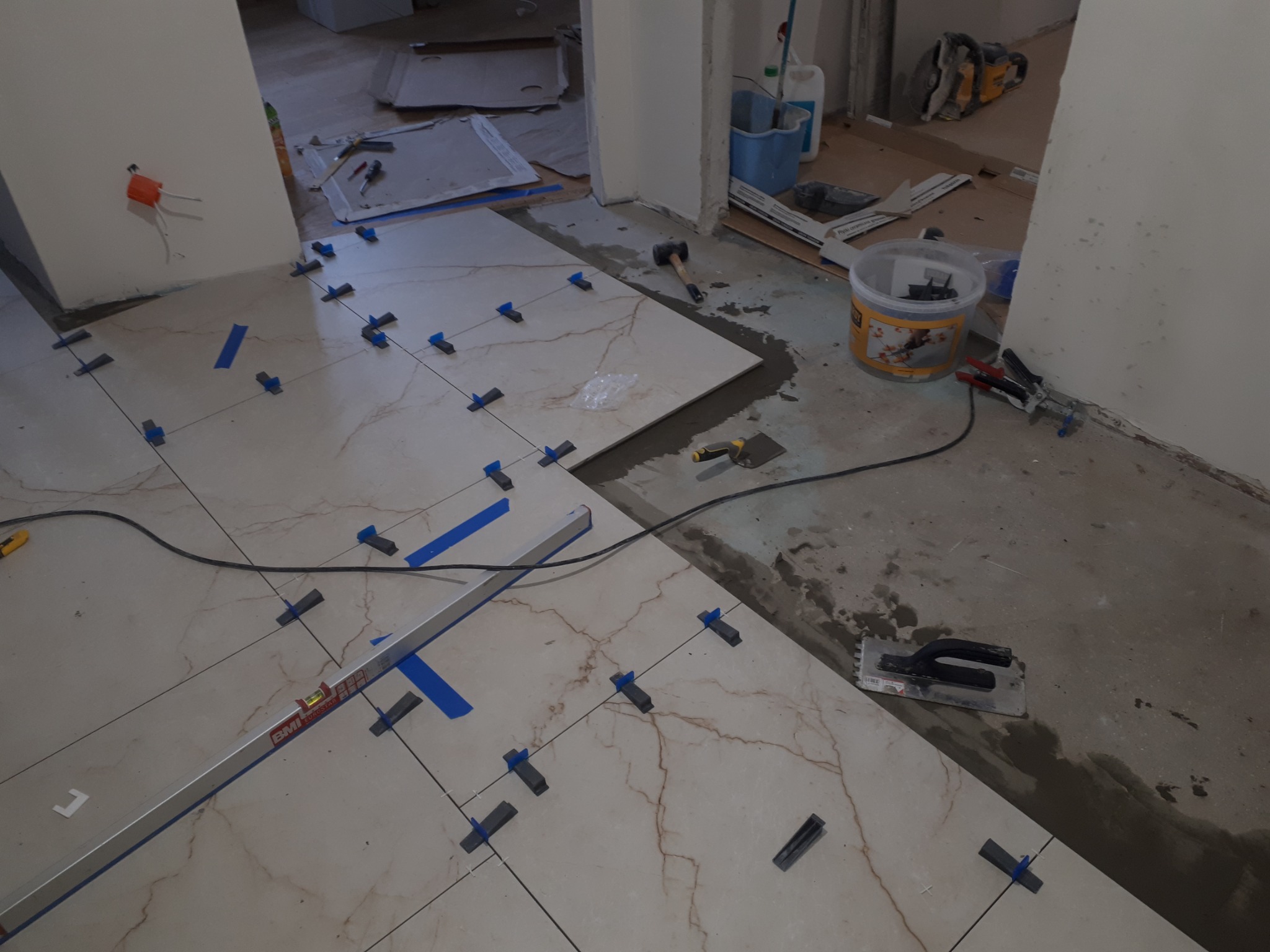 Układanie płytek podłogowych z użyciem poziomicy BMI, krzyżyków dystansowych i taśmy malarskiej; widoczny klej, kielnia i młotek gumowy.