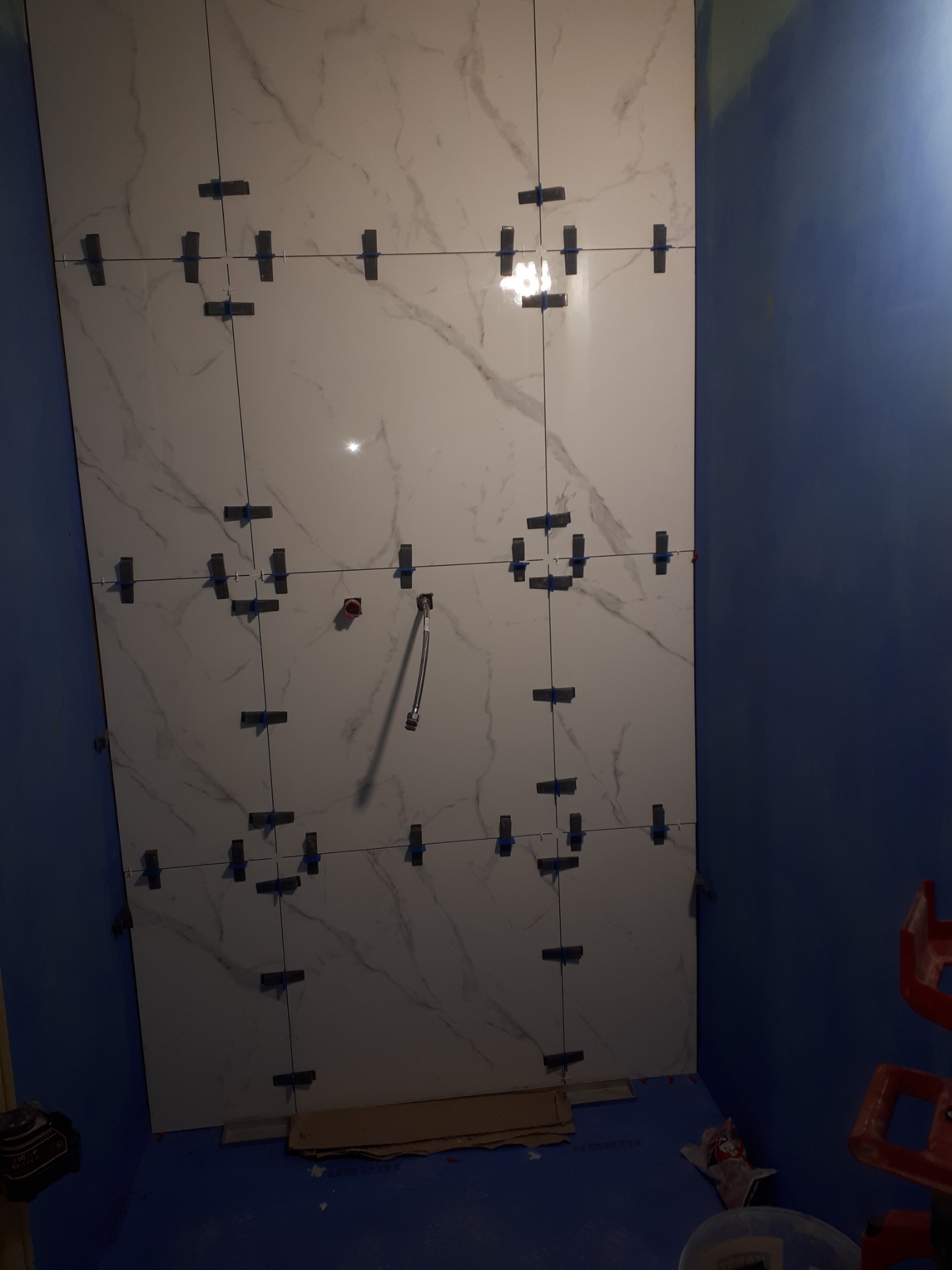 Ściana łazienki w trakcie układania płytek imitujących marmur, z widocznymi krzyżykami dystansowymi i wystającą rurką instalacyjną, niebieskie ściany i podłoga zabezpieczona folią.