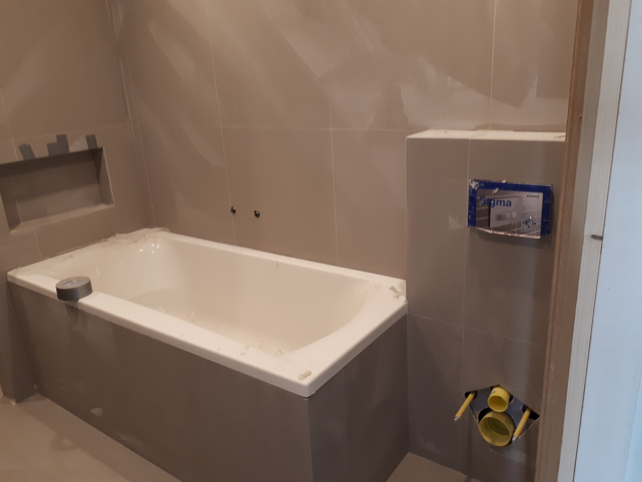 Remont łazienki w trakcie: biała wanna obudowana szarą płytką, widoczne przyłącza kanalizacyjne i zabudowa pod stelaż WC z zamontowanym modułem spłukującym Geberit Sigma.