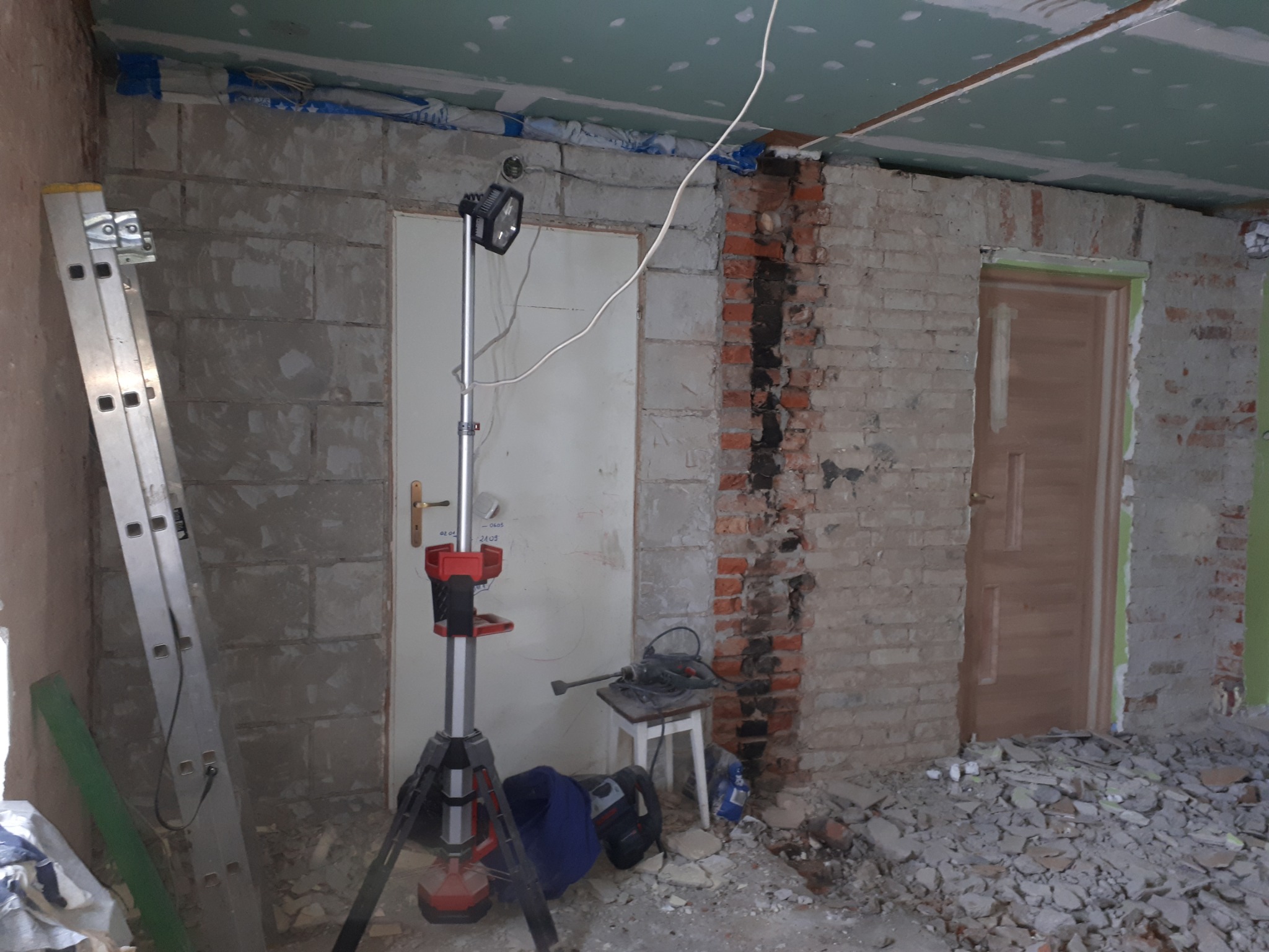 Wnętrze w trakcie prac: odsłonięta cegła, surowe ściany z bloków, częściowo położone płyty gipsowo-kartonowe na suficie, stojąca lampa budowlana na trójnogu, drabina aluminiowa oparta o ścianę...