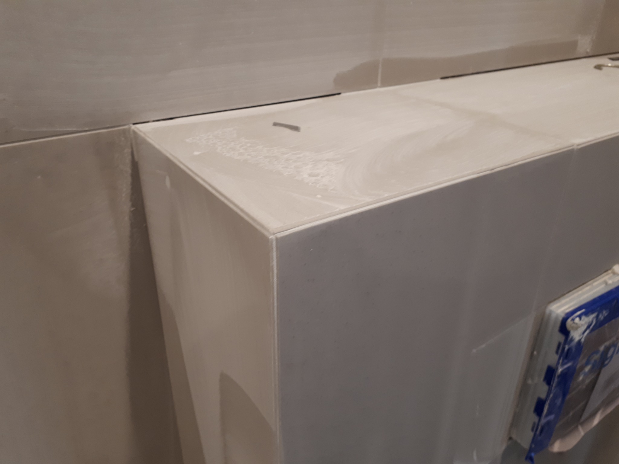 Szare płytki ceramiczne na zabudowie stelaża WC z widocznymi śladami po fugowaniu i drobnym zabrudzeniem na górnej powierzchni. Użyto poziomicy do precyzyjnego ułożenia płytek.