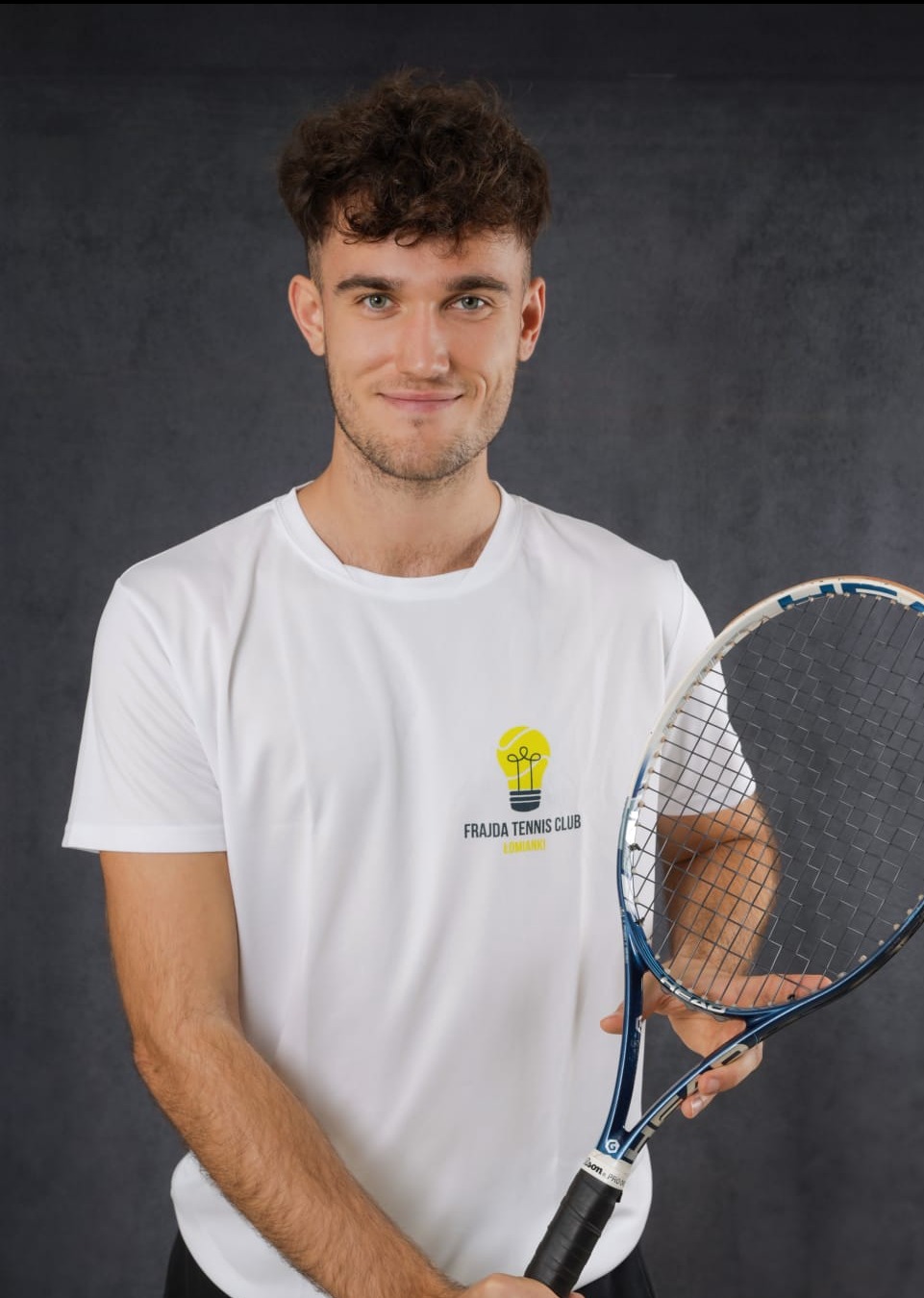 Uśmiechnięty młody mężczyzna w białej koszulce z logo Frajda Tennis Club trzyma rakietę tenisową na tle ciemnoszarego tła.