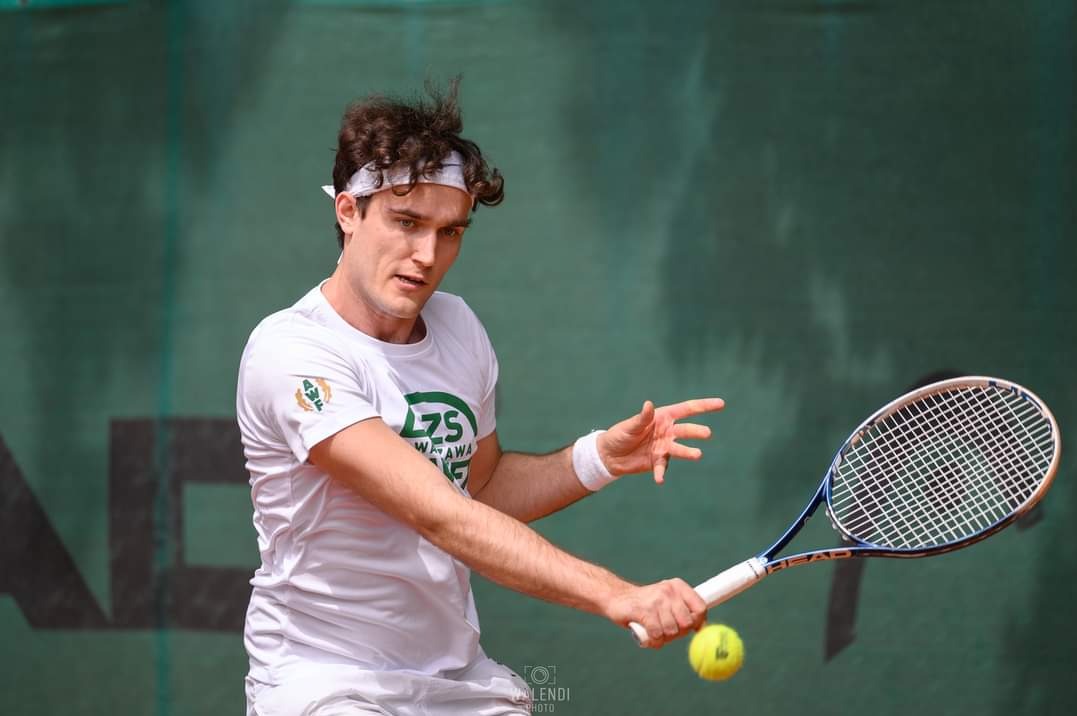 Młody mężczyzna z opaską na głowie, w trakcie uderzenia piłki tenisowej rakietą na tle kortu ziemnego.