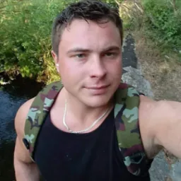 Młody mężczyzna z plecakiem moro, stojący obok strumienia, z widoczną srebrną biżuterią na szyi i w uchu.