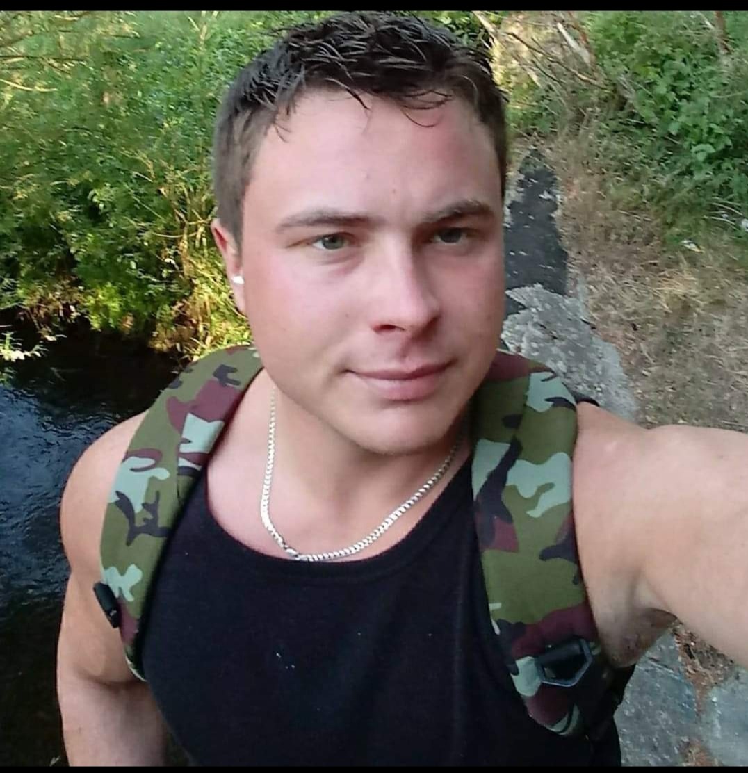 Młody mężczyzna z plecakiem moro, stojący obok strumienia, z widoczną srebrną biżuterią na szyi i w uchu.