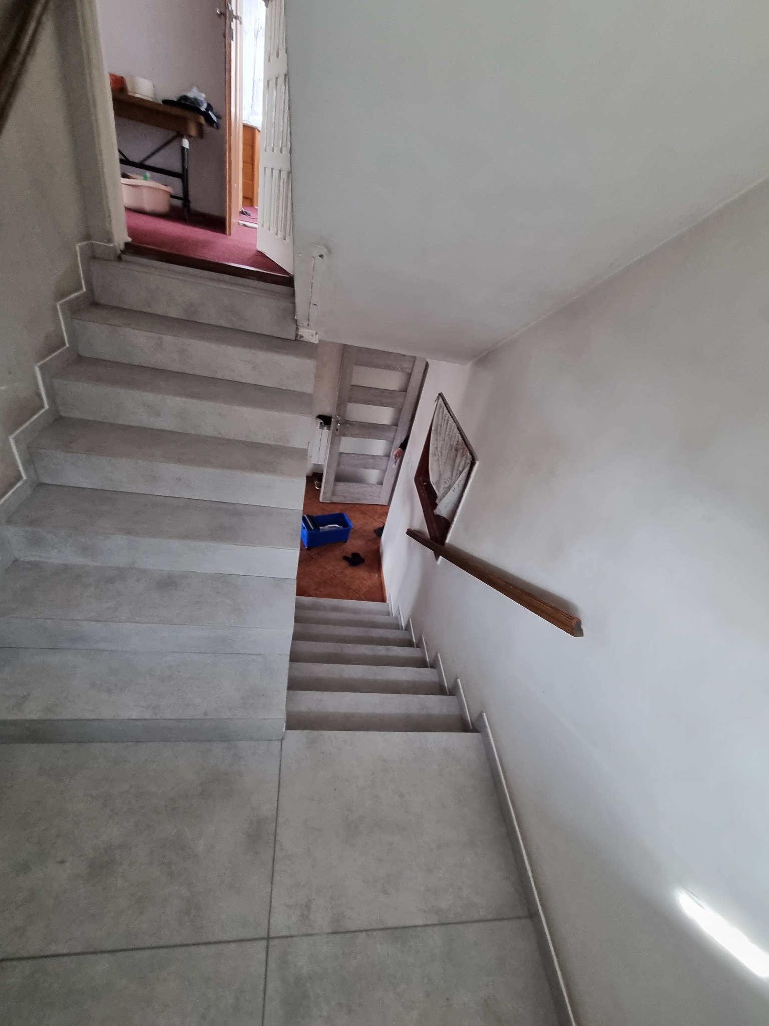 Widok z góry na schody wyłożone szarą płytką, z drewnianą poręczą po prawej stronie i niebieskim pojemnikiem na dole schodów, prowadzące do pomieszczenia z czerwonym dywanem i białymi drzwiami.
