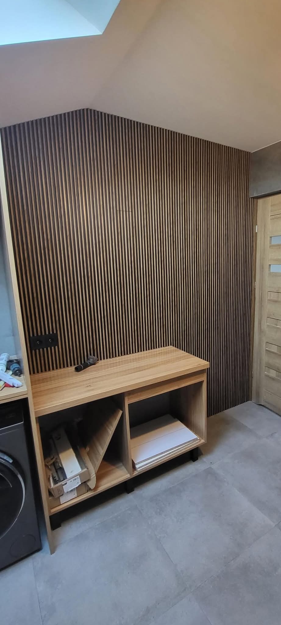 Ściana w łazience wykończona panelami lamelowymi w odcieniach brązu, z drewnianą szafką pod spodem i widocznym gniazdkiem elektrycznym. Na szafce leży mikrofon. Podłoga wyłożona szarymi płytkami.