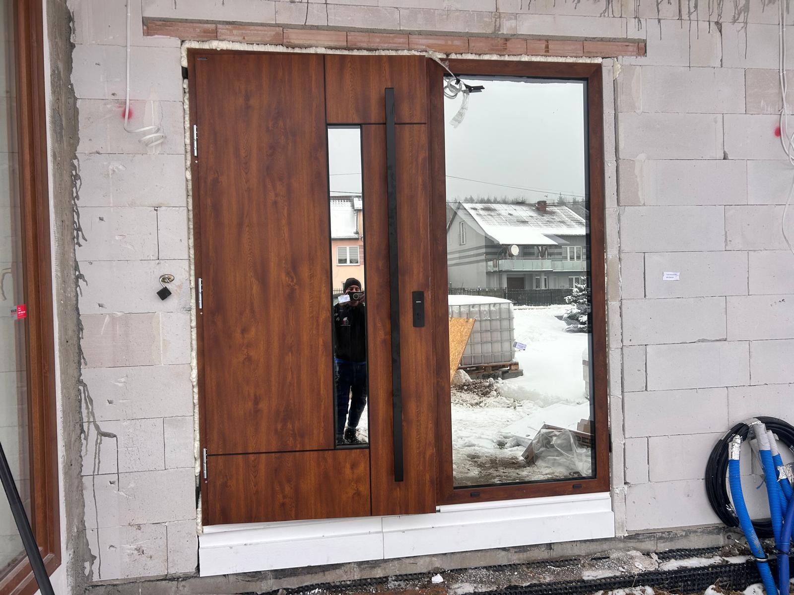 Montaż nowoczesnych drzwi wejściowych z przeszkleniem w Miedzianej Górze, widoczny surowy mur i ośnieżony krajobraz w odbiciu szyby.