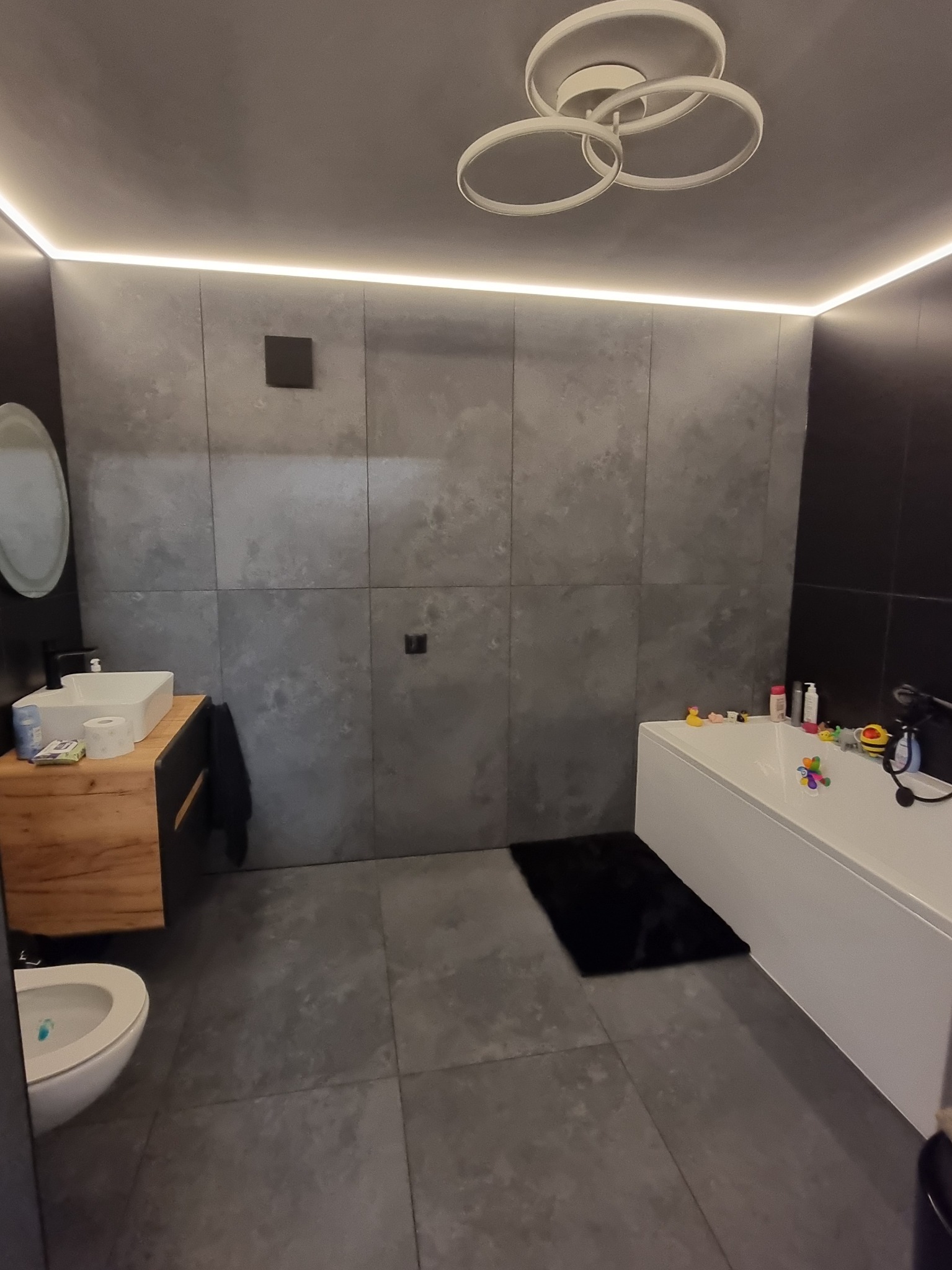 Nowoczesna łazienka z szarą, kwadratową glazurą ścienną, podświetleniem LED nad szafką i sufitem, oraz okrągłą lampą sufitową z trzema pierścieniami. Widoczna umywalka na drewnianej szafce, toaleta...