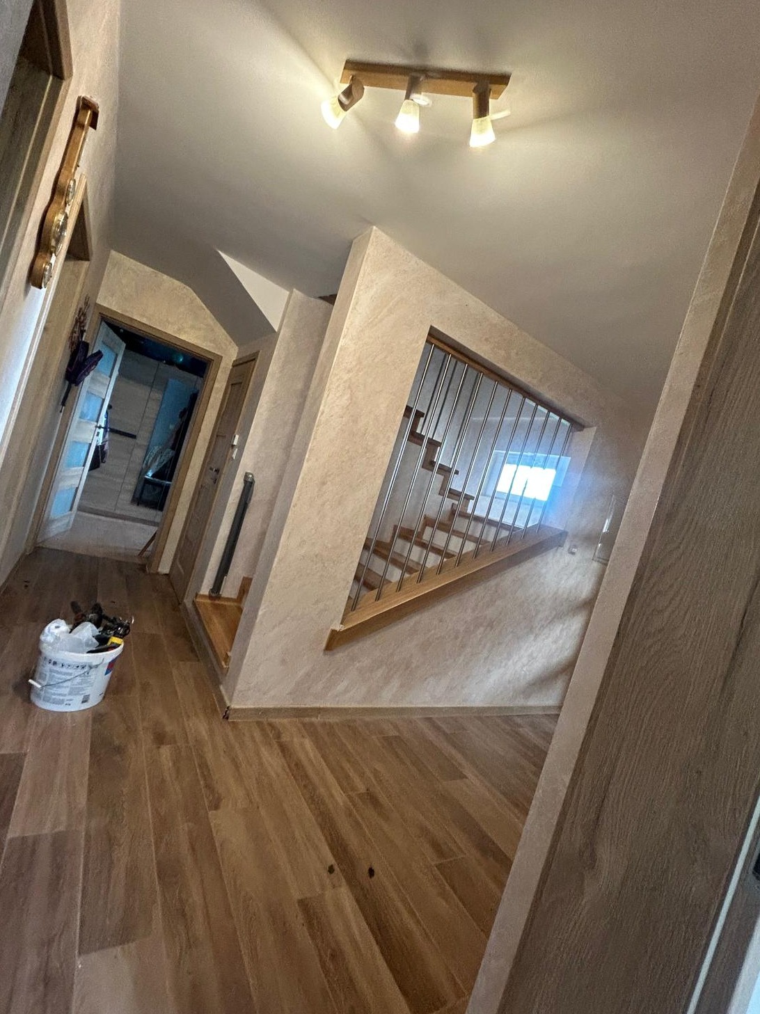 Korytarz po remoncie z drewnianą podłogą, widoczne schody z metalową barierką, drzwi i wiadro z narzędziami malarskimi w rogu.