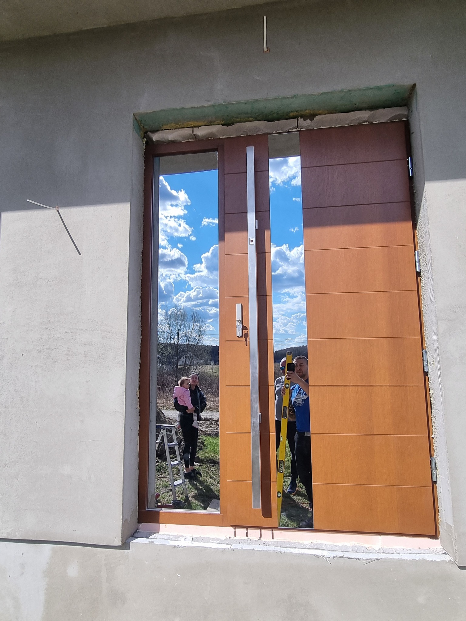 Montaż nowoczesnych, brązowych drzwi zewnętrznych z pionowymi przeszkleniami, widoczne poziomowanie z użyciem poziomicy, odbicie montażystów i krajobrazu w szybach.