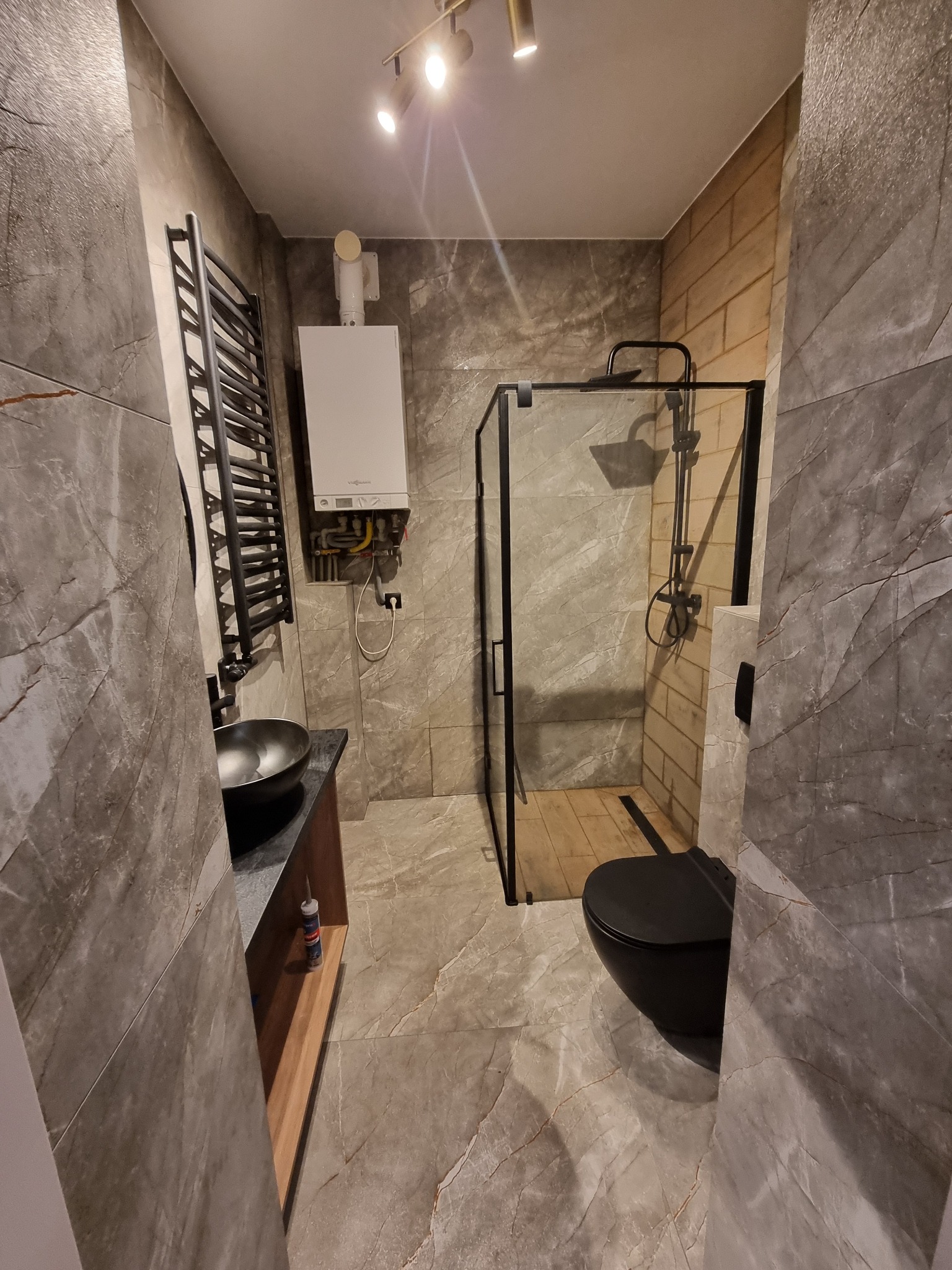 Nowoczesna łazienka z płytkami imitującymi marmur, czarną armaturą i kabiną prysznicową typu walk-in. Widoczny piec gazowy i grzejnik drabinkowy.