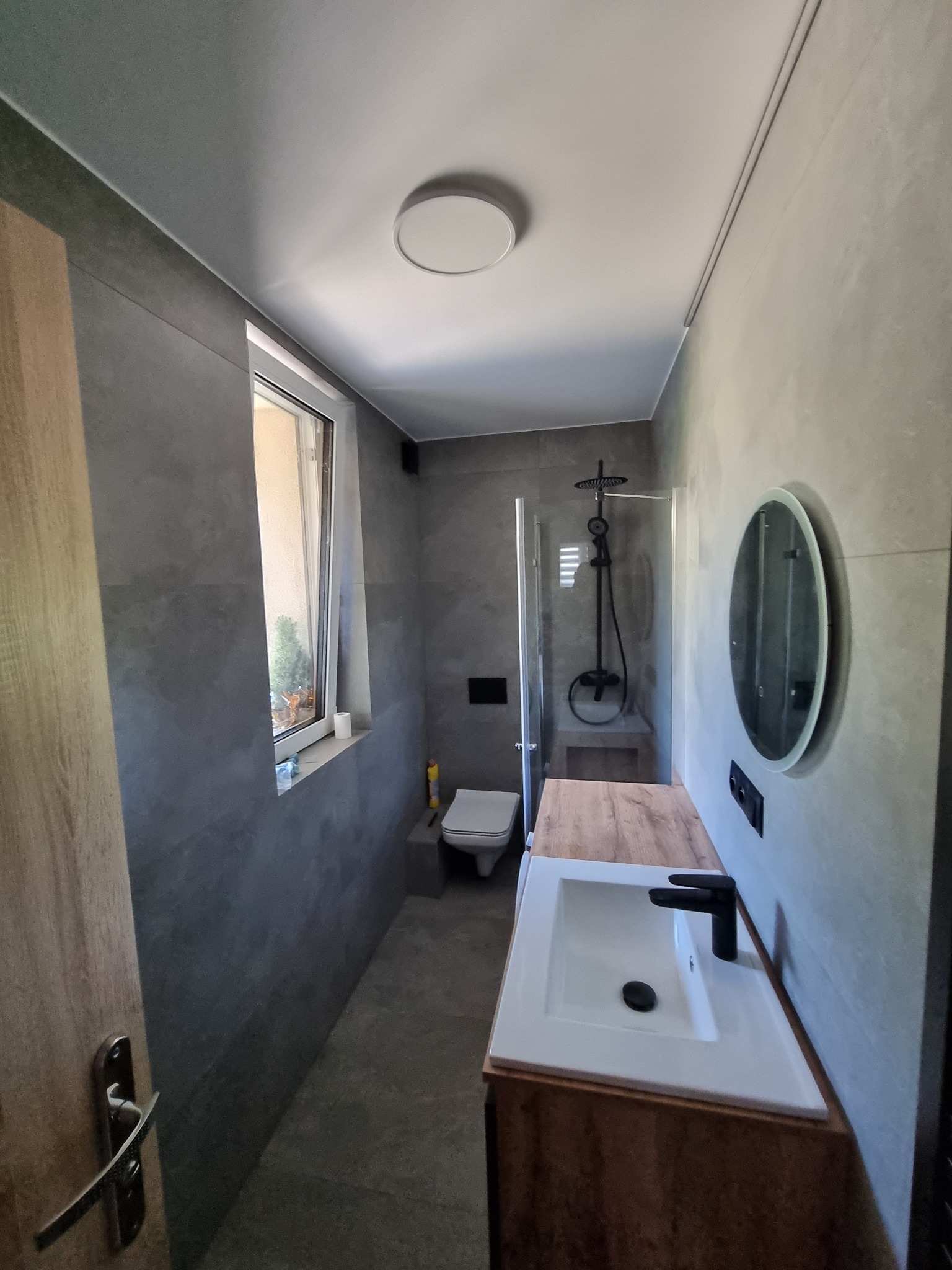 Nowoczesna łazienka w szarościach z drewnianym blatem pod umywalką i okrągłym lustrem. Kabina prysznicowa ze szklaną ścianką. Widok z perspektywy wejścia.