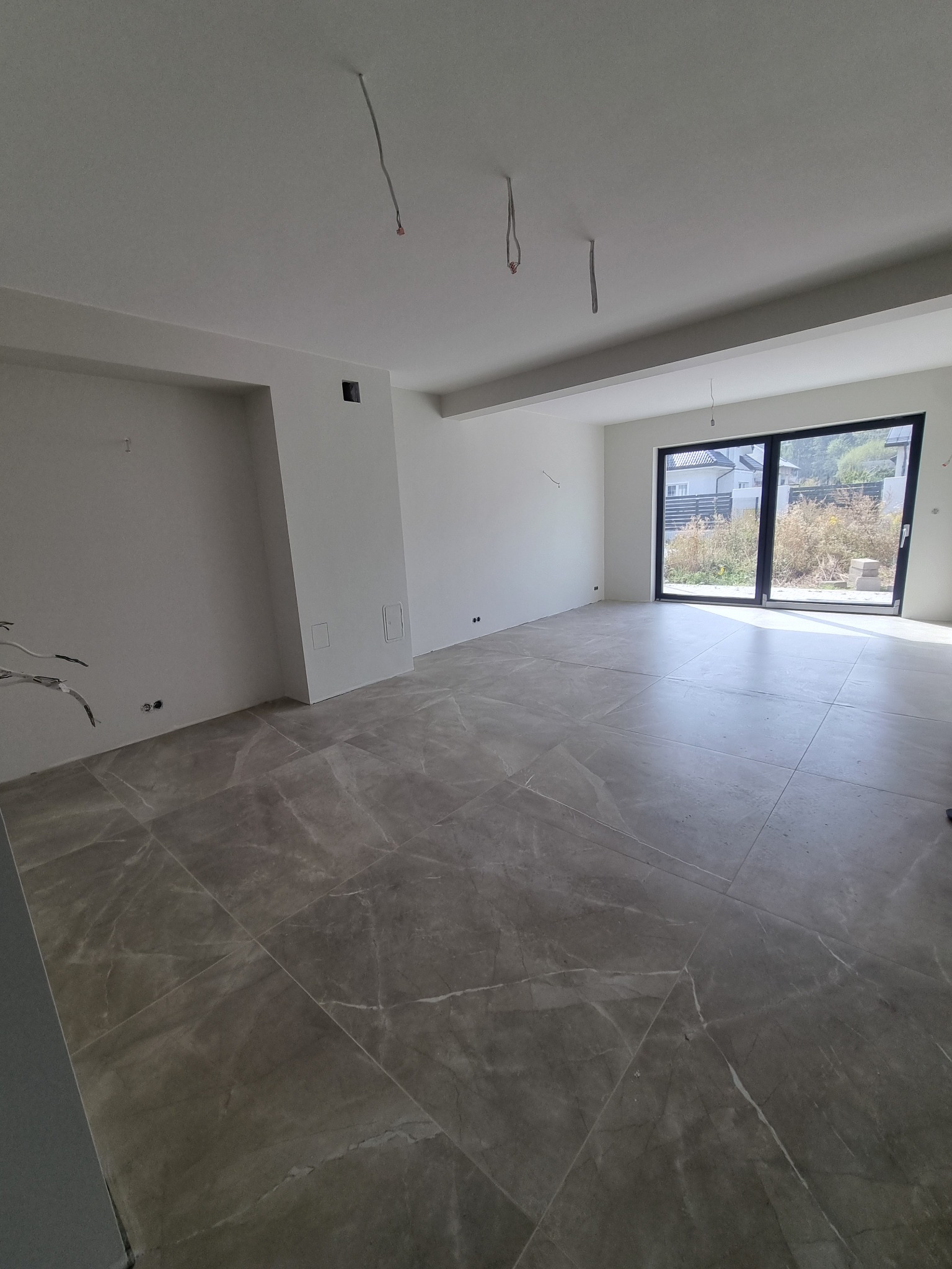 Jasne wnętrze z szarymi płytkami imitującymi marmur, świeżo po wykończeniu. Wiszące przewody elektryczne i widok na zewnątrz przez duże okno balkonowe.