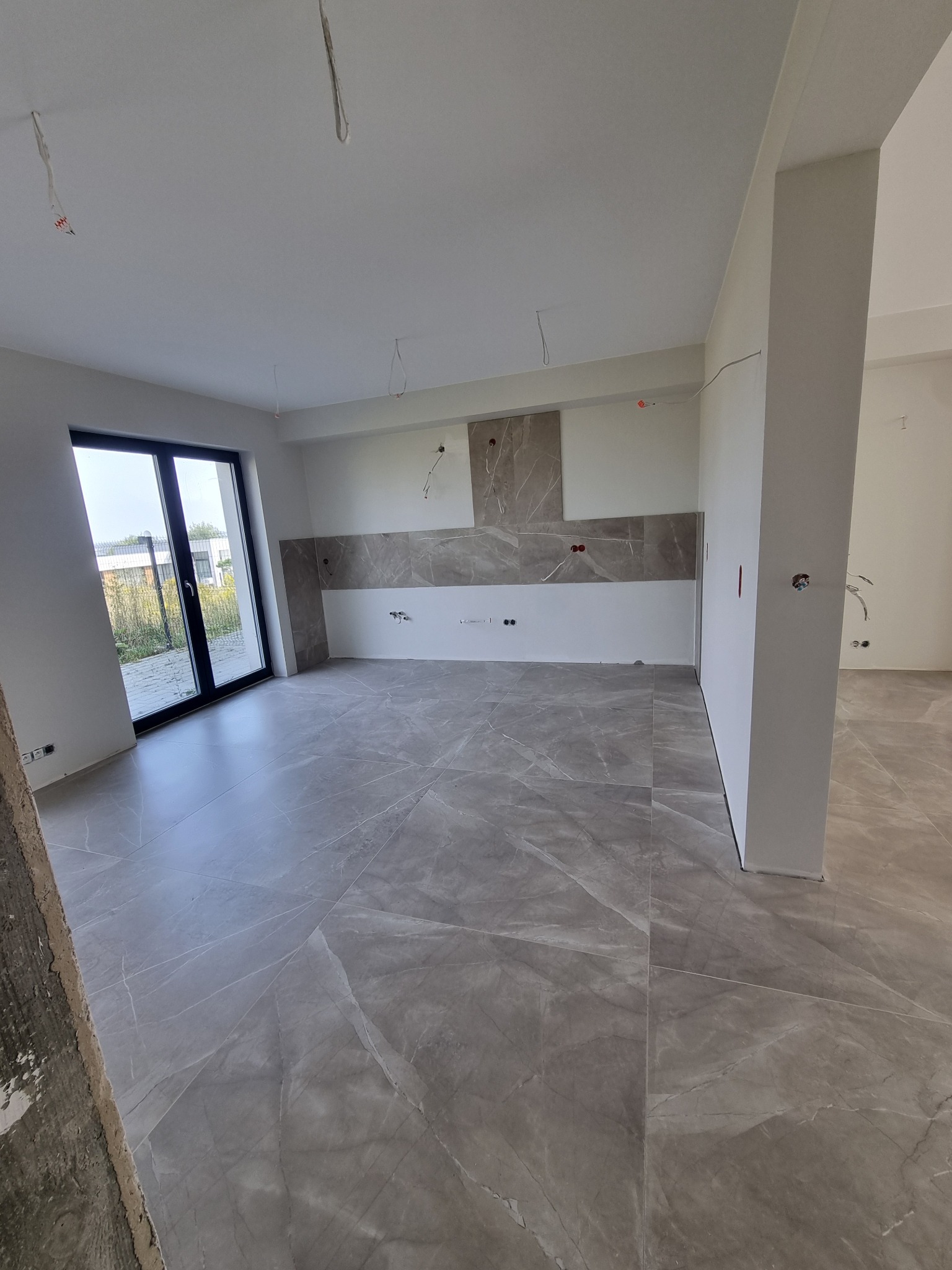 Wykończone wnętrze: szara, marmurkowa podłoga z płytek, częściowo wykończona ściana z szarymi płytkami. Widoczne niedokończone instalacje elektryczne.