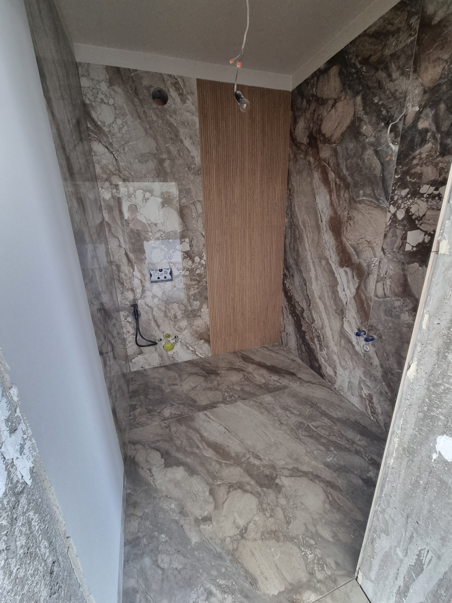 Łazienka w trakcie wykańczania: marmurowe płytki na ścianach i podłodze, drewnopodobna okładzina ścienna, widoczne instalacje. Ujęcie z perspektywy wejścia.