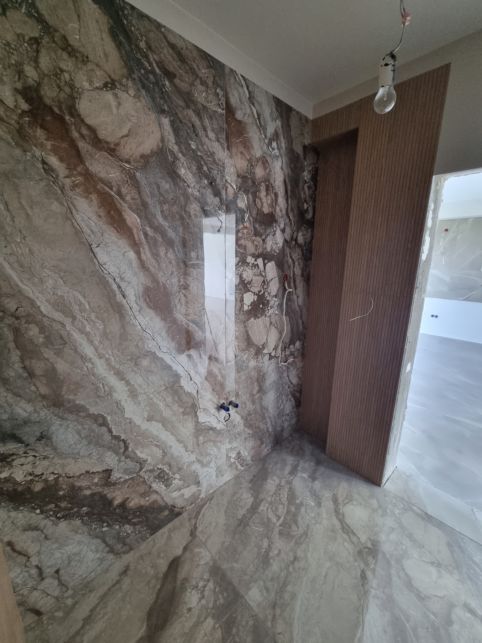 Ściana i podłoga wyłożone płytkami imitującymi marmur w łazience. Widoczne przewody elektryczne i surowa żarówka. Wykończenie w trakcie realizacji.