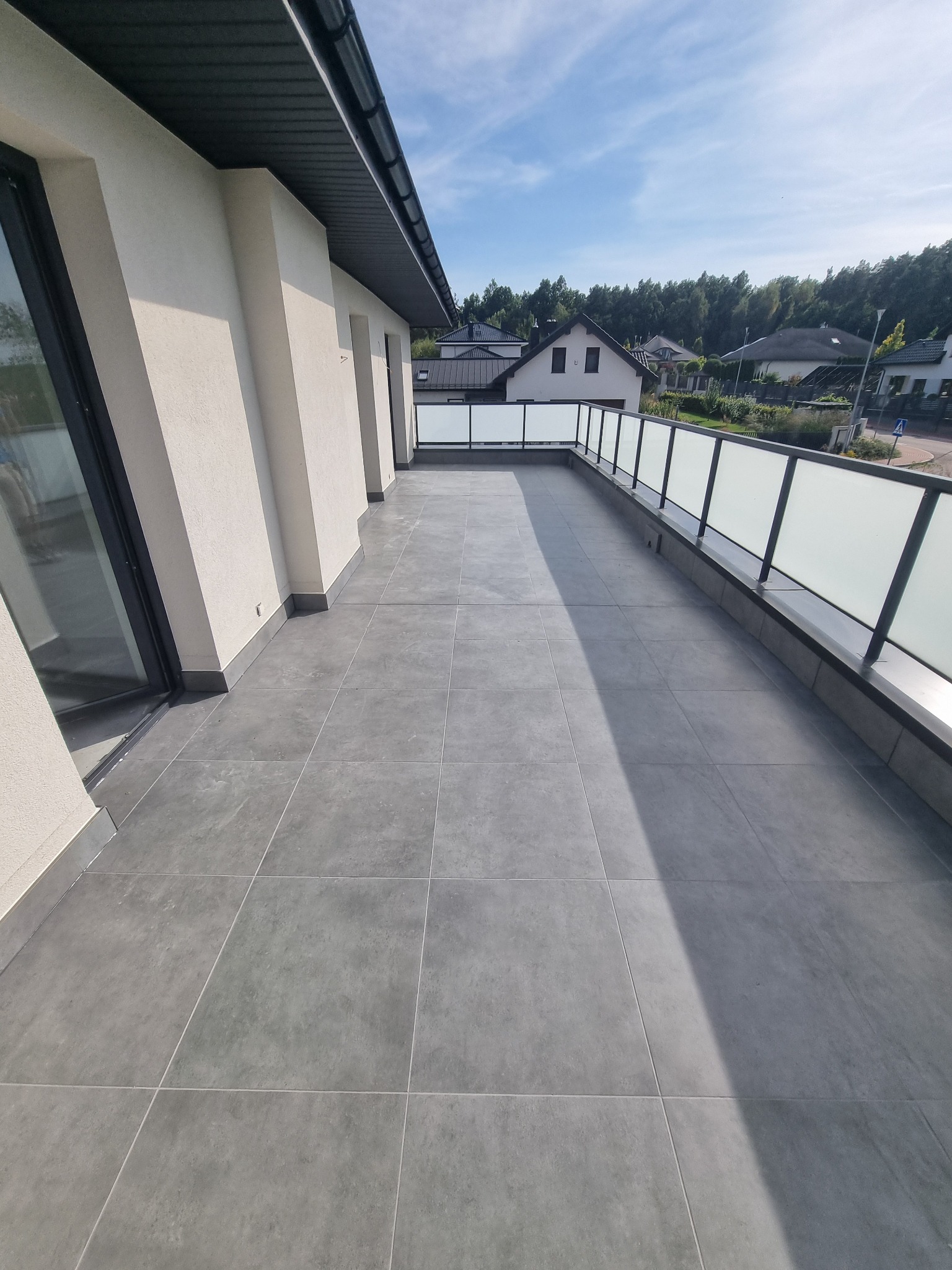 Wykończony balkon z szarą, kwadratową terakotą i szklaną balustradą. Widok na okoliczne domy i zieleń w słoneczny dzień. Minimalistyczny styl.