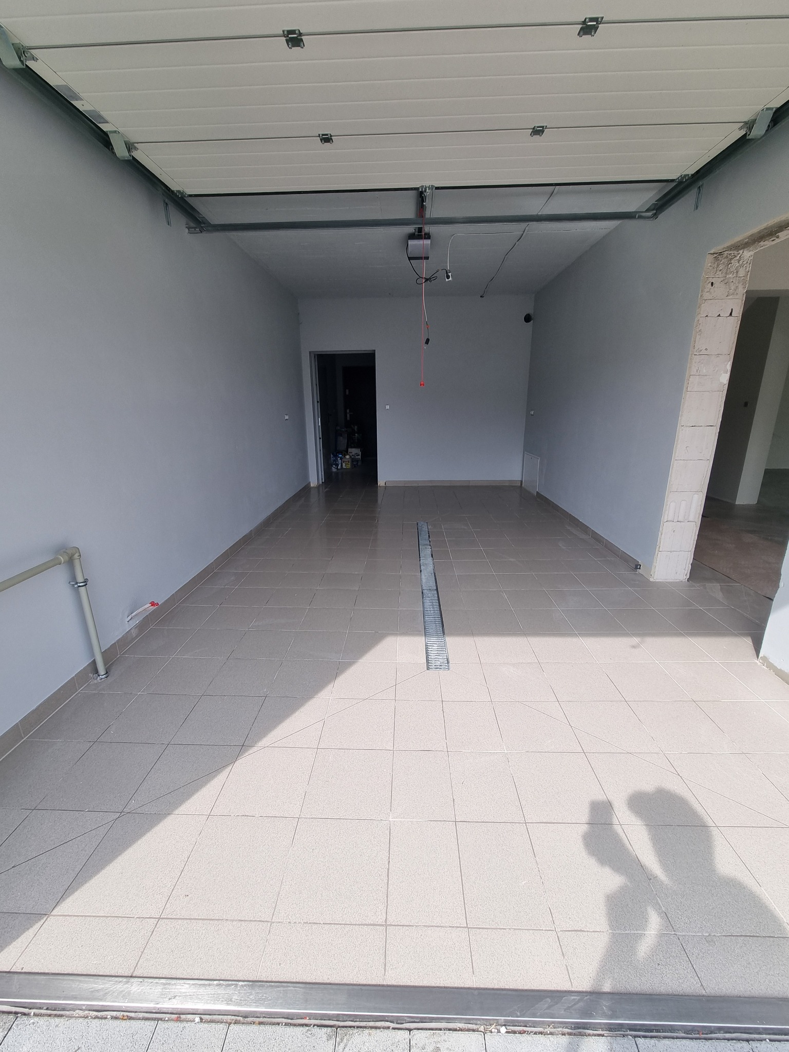 Wykończone wnętrze garażu z beżowymi płytkami podłogowymi i szarymi ścianami, widoczny odpływ liniowy. W tle otwarte drzwi do innego pomieszczenia.