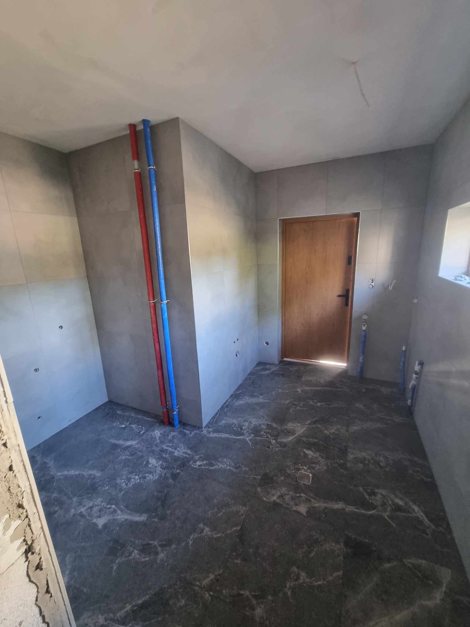 Szara łazienka w trakcie wykończenia: płytki na ścianach i podłodze, widoczne rury instalacji wodnej. Drzwi wejściowe w tle, surowy stan.