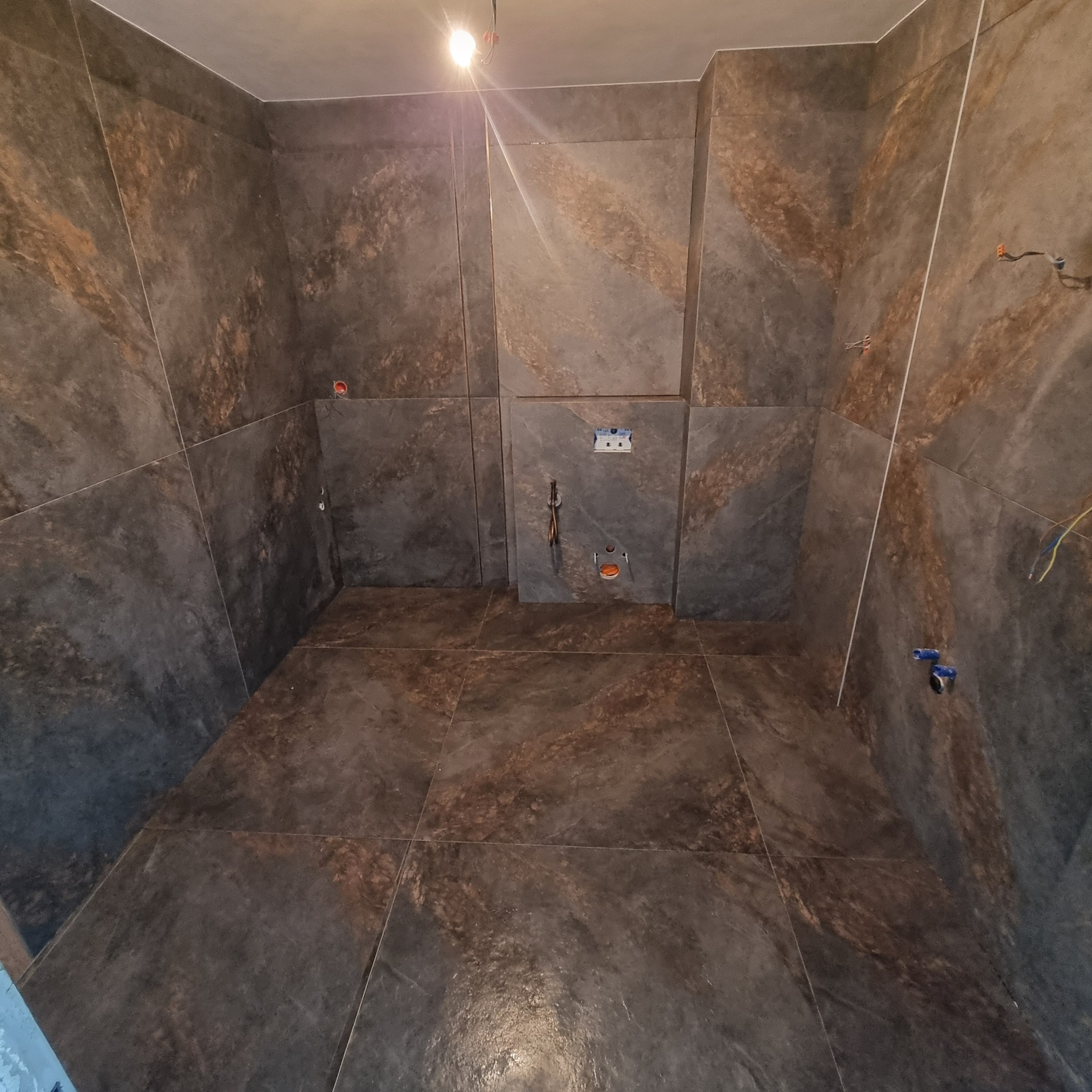 Wykończona łazienka z ciemnymi, marmurkowymi płytkami na ścianach i podłodze. Widoczne gniazdka elektryczne i instalacja wodna przed montażem armatury.