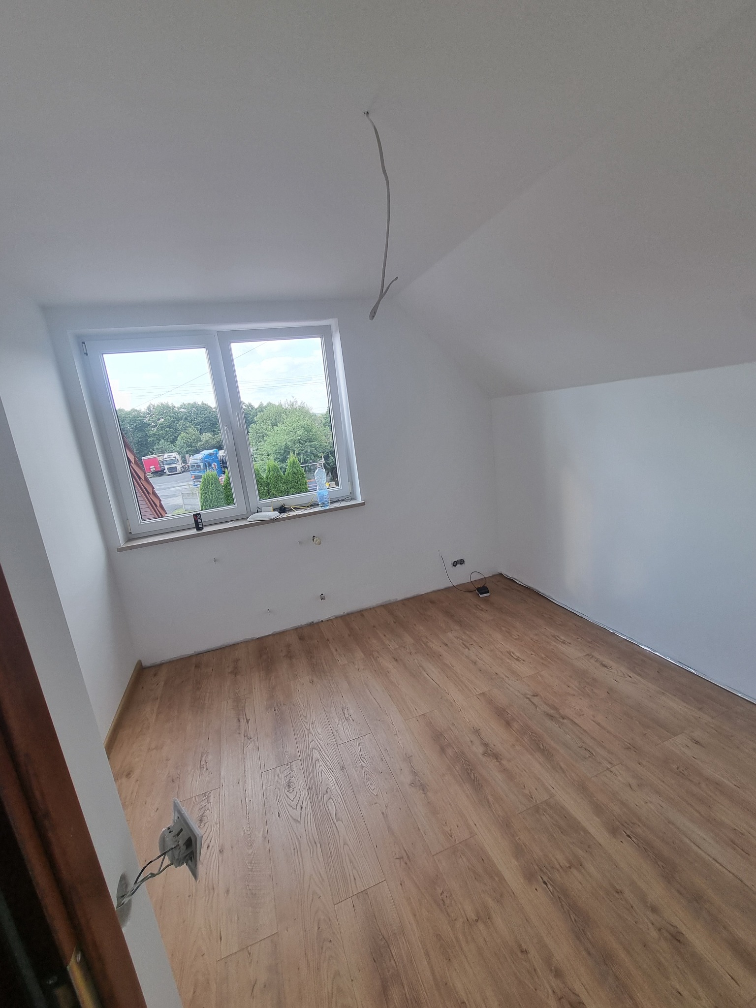 Pokój na poddaszu z ułożonymi panelami podłogowymi w odcieniach brązu. Widoczne białe ściany, okno z widokiem na zewnątrz i wystający kabel ze ściany.