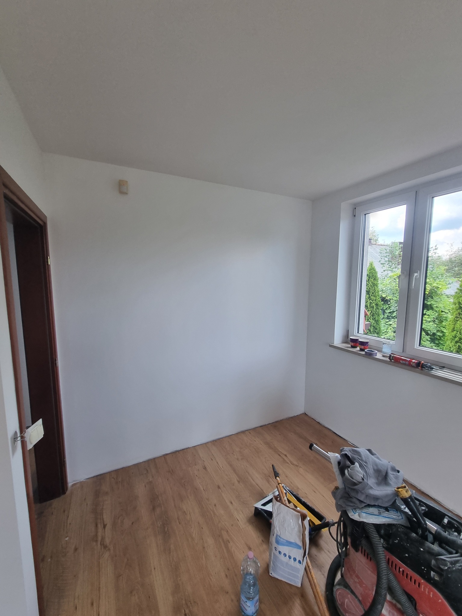 Pomalowane na biało wnętrze pokoju z drewnianą podłogą. Widoczne narzędzia malarskie, okno z widokiem na zieleń i brązowe drzwi.