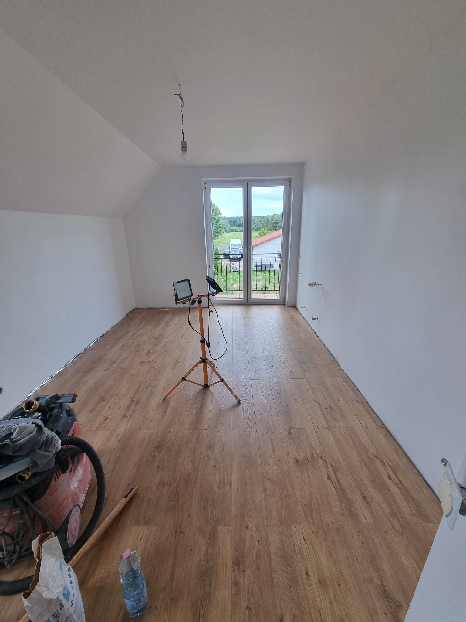 Poddasze po remoncie: ułożone panele podłogowe, białe ściany, okno balkonowe z widokiem na zieleń, lampa robocza na statywie, narzędzia w rogu pokoju.