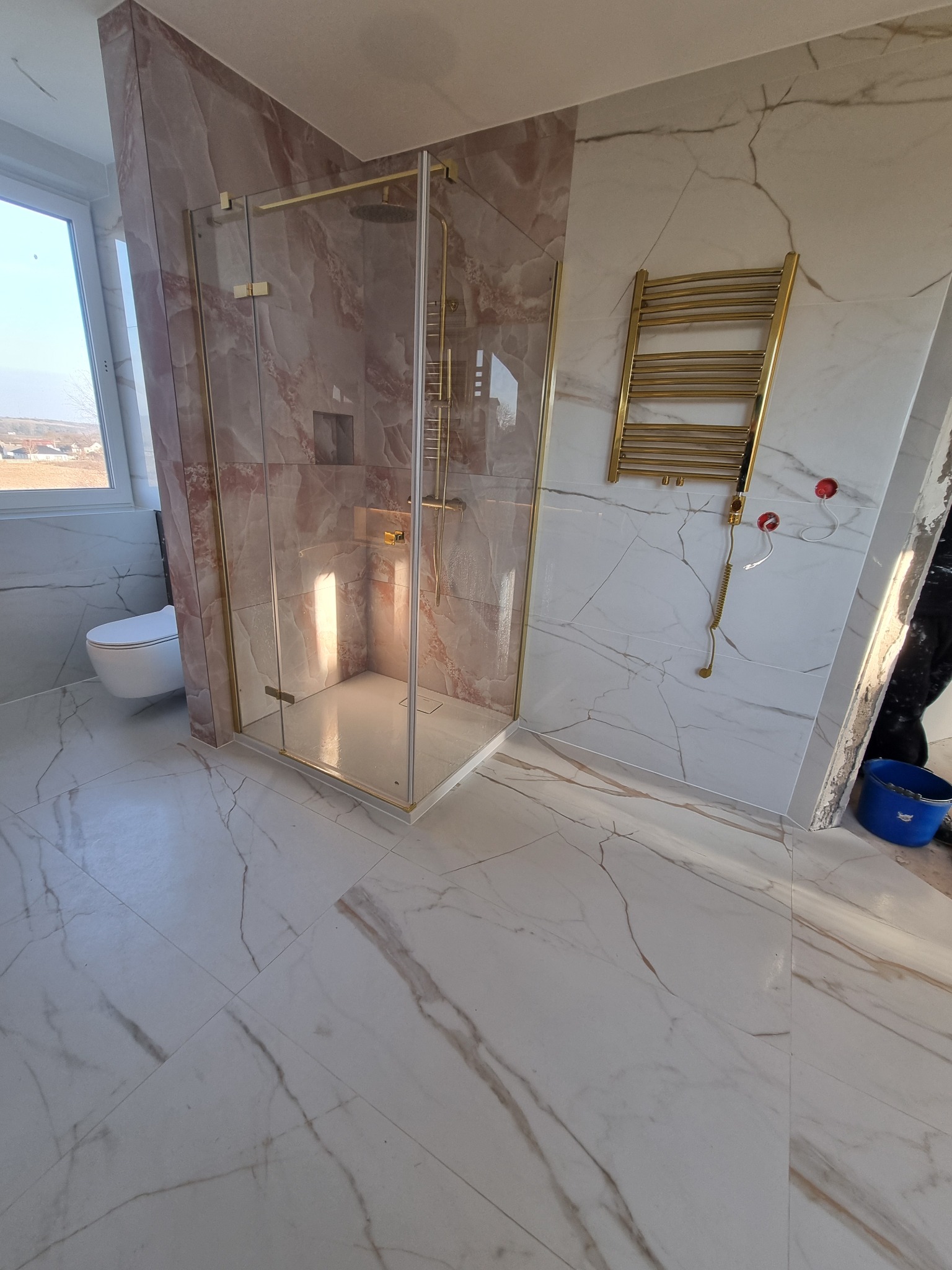 Wykończona łazienka z kabiną prysznicową w odcieniach beżu i złota, marmurowe płytki na ścianach i podłodze, grzejnik drabinkowy w złotym kolorze.