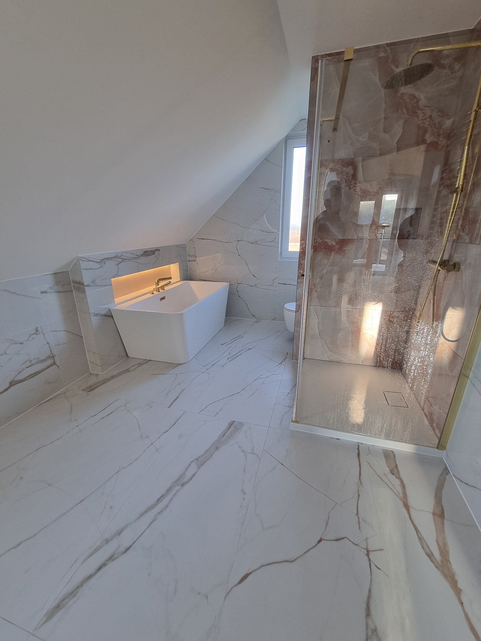 Nowoczesna łazienka na poddaszu z marmurowymi płytkami, białą wanną pod skosem i kabiną prysznicową z elementami w kolorze złota. Minimalistyczny design.