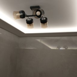 Grzegorz Karbowiak - Czarny plafon z trzema kloszami w odcieniach brązu, zamontowany na białym suficie z oświetleniem LED, widoczna ściana wykończona płytami imitującymi marmur.